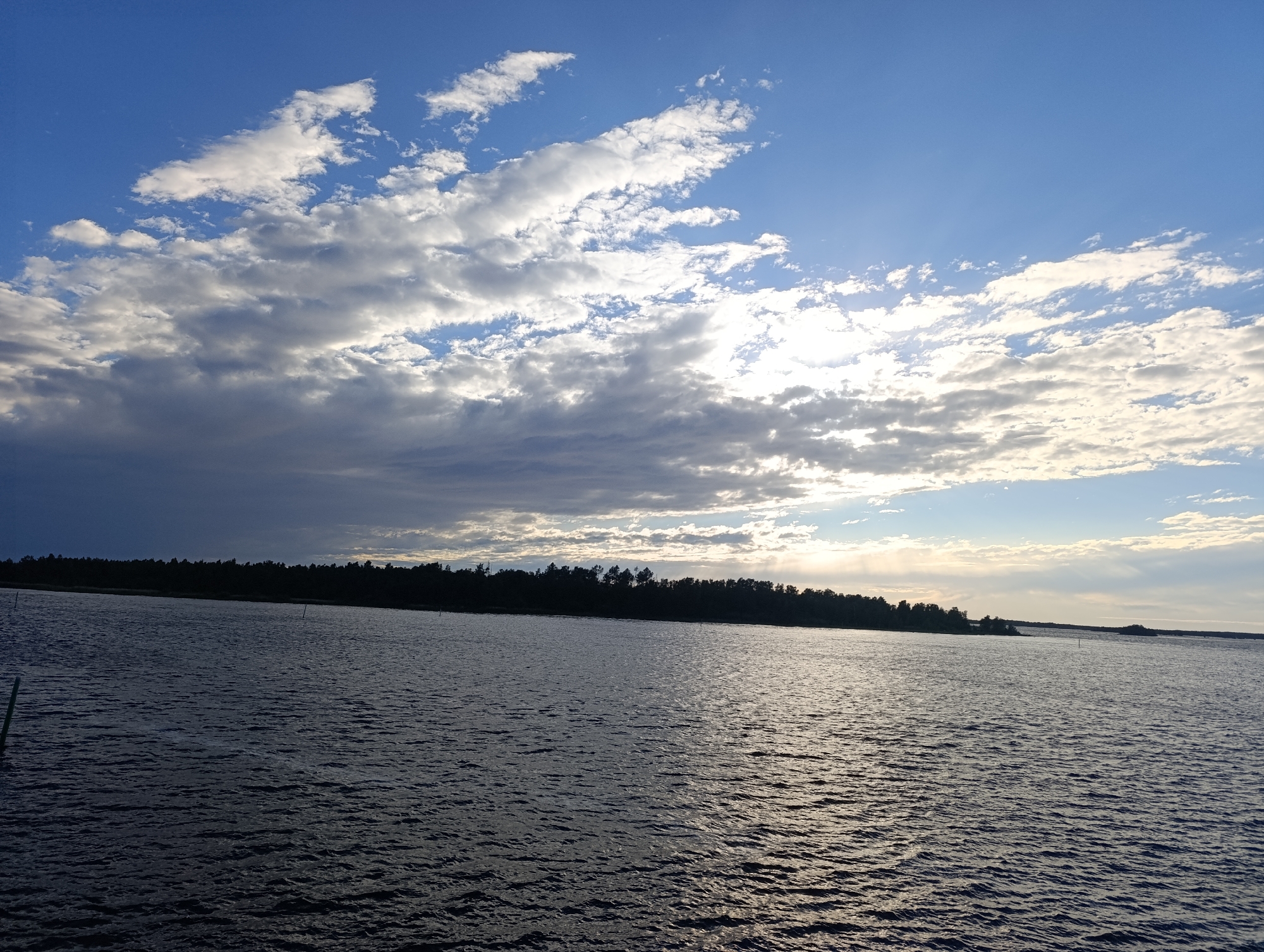 The image depicts a wide expanse of dark, rippling water under a dramatic sky. The sky is a mix of bright blue near the horizon, with scattered white and gray clouds. Sunbeams are visible breaking through the clouds, creating a bright, slightly hazy effect in the upper right quadrant of the image. A dark, silhouetted treeline runs horizontally across the middle ground, separating the water from the sky. The treeline suggests a forested shoreline. The water's surface is textured with small waves, reflecting the light from the sky. There are a few indistinct vertical objects in the water near the left edge and in the distance, possibly markers or small poles. The scene appears to be a natural landscape, likely a lake or a bay, given the presence of a treeline and the broad body of water. The lighting suggests it is either sunrise or sunset. The image is a landscape photograph, capturing a serene natural environment. This scene is characteristic of the coastal areas and numerous lakes found around Jakobstad, Finland, particularly in the archipelago or along the mainland shores.