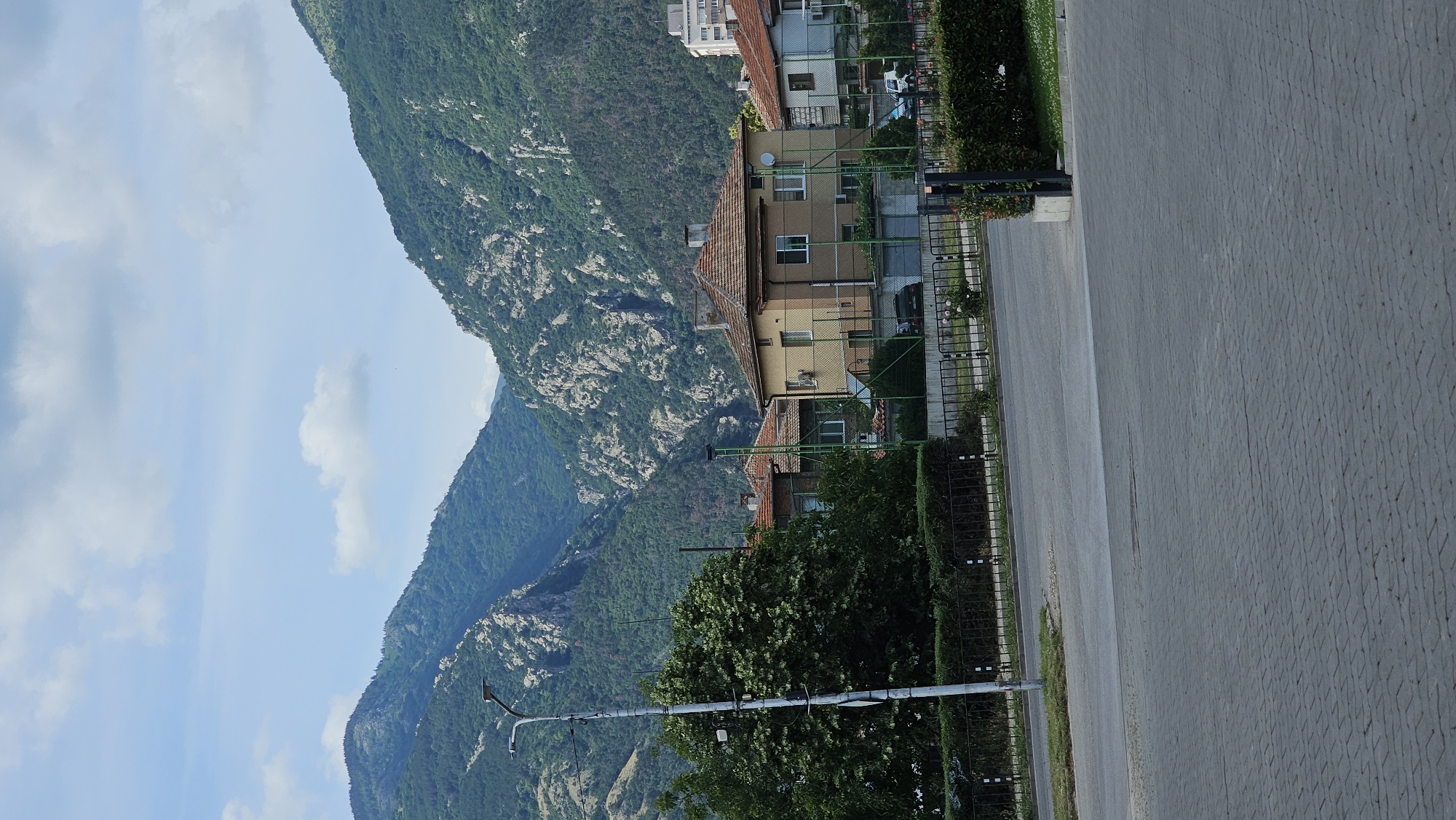 This is a vertical, eye-level, full shot of a residential area in Karlovo, Bulgaria, framed by a dramatic mountain landscape. In the foreground, a paved road with cobblestone paving is visible. Beyond the road, a black metal fence delineates the property line of several houses. The houses are two-story structures with terracotta-tiled roofs and light-colored facades. A large, leafy green tree stands to the left of the houses. A tall, gray street lamp with multiple lights is positioned in front of the tree. The middle ground features the houses and a lush green lawn with manicured hedges. In the background, imposing mountains rise steeply, covered in dense green vegetation and rocky outcrops. The sky is partly cloudy with scattered white clouds against a light blue backdrop. No people are visible in the image. The scene appears to be a quiet, suburban setting at the foot of a mountain range.