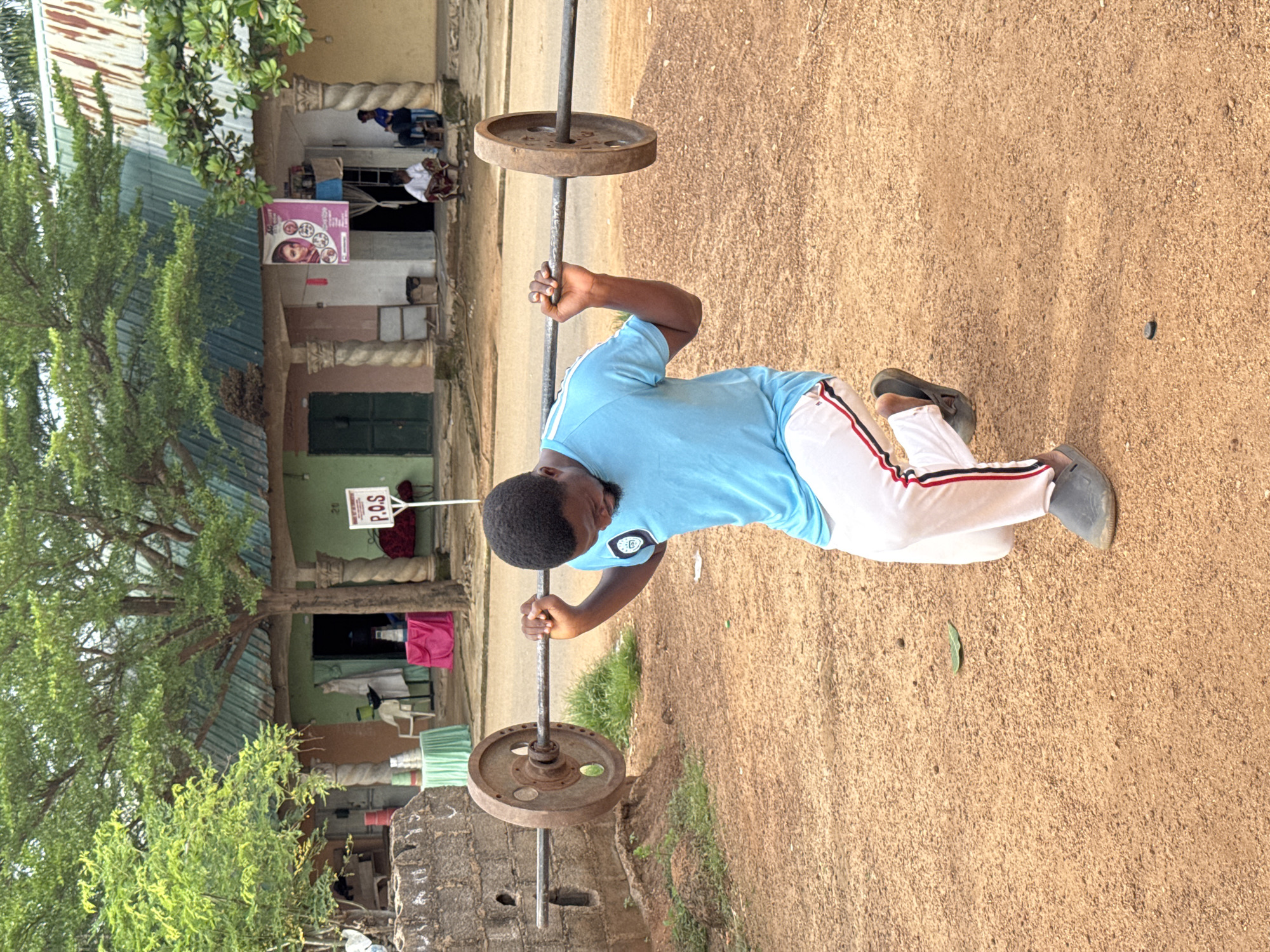 A man is performing a weighted lunge with a barbell outdoors in front of a building in Akwanga, Nigeria. The man, who appears to be of African descent, is wearing a light blue t-shirt with a patch on the left sleeve and white pants with red and black stripes down the sides. He is in a lunge position with his left leg forward and bent at the knee, and his right leg extended backward. He is holding a barbell across his upper back, with the weight plates visible on either side.
The ground is covered in reddish-brown dirt and gravel. Behind the man, a one-story building with a green corrugated metal roof is visible. The building has several entrances and windows, some of which are open. There are decorative pillars in front of the building. A sign that reads "P.O.S" is mounted on a post in front of the building. Trees with green foliage are present in the foreground and behind the building. The overall scene suggests an informal outdoor workout setting.