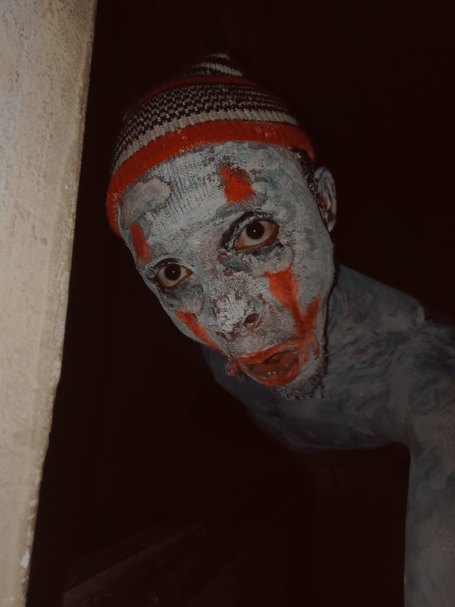 A close-up, dimly lit portrait features a person with their face and visible upper body covered in light blue or grey body paint. They wear a red, white, and black striped knit beanie hat. Streaks of bright red paint are applied to the face, resembling blood or ritualistic markings, particularly around the wide-open eyes, the bridge of the nose, and below the slightly agape mouth. The texture of the body paint appears somewhat cracked on the skin. The subject's dark eyes gaze intently and directly at the viewer, conveying an intense and somewhat unsettling expression.
The person is partially obscured, positioned as if peeking from behind a light-colored, possibly stuccoed wall or doorway that occupies the left side of the frame. The background behind the subject is intensely dark, suggesting an unlit interior space or a night-time setting, which dramatically highlights the painted features. The overall atmosphere of the scene is eerie and mysterious, evoking feelings of unease or intrigue. This could potentially be part of a performance, a cultural ceremony, or a Halloween-themed portrayal. No visible text is present in the image.