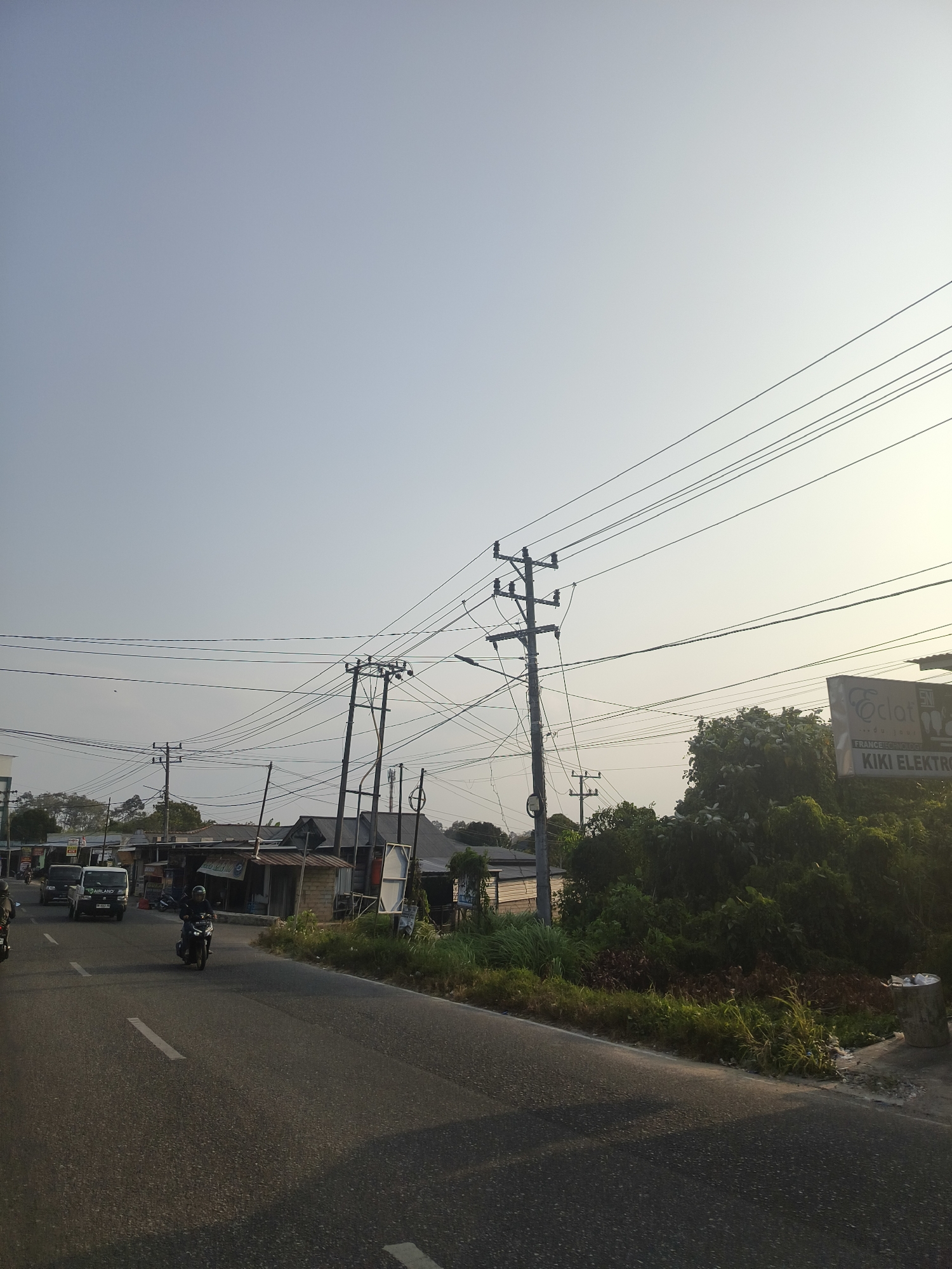 The image depicts a street scene in Pangkalpinang, Indonesia, likely during the late afternoon given the soft, warm lighting. The perspective is from the side of a paved road, which occupies the lower portion of the frame. The road has white lane markings. On the left side of the road, several vehicles are visible, including a black car and a motorbike with a rider. Further down the road, small buildings and shops line the street, appearing to be a commercial or residential area with some structures in disrepair. Numerous utility poles with a dense network of electrical wires crisscross the sky, a prominent feature of the urban landscape.
To the right of the road, there is a thick growth of trees and vegetation. A signpost with the text "Oclat" and "KIKI ELEKTRO" is partially visible amidst the greenery, suggesting a local business. The sky is a pale, hazy blue, indicating clear weather. The overall impression is of a typical street in a developing urban area, characterized by its infrastructure, local commerce, and natural elements.