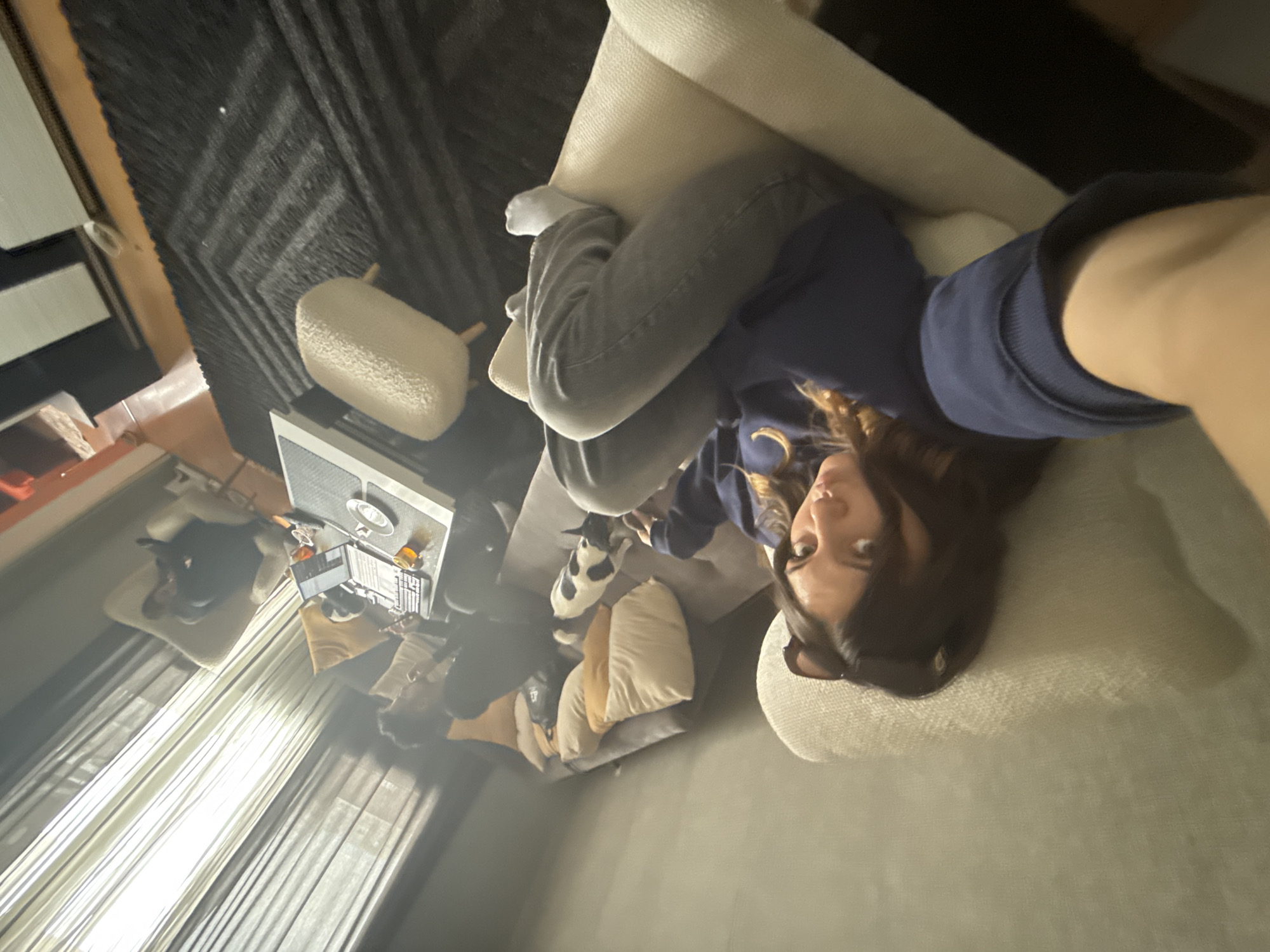 A selfie taken inside a room in Sofia, Bulgaria, shows a young woman lying on a light-colored sofa wearing gray pants and a blue hooded sweatshirt. A white dog with black spots lies near her on the sofa. To the left is a coffee table with a laptop, a plate, and a glass. Behind the sofa are pillows and a seated person. Visible through a window are curtains and a light-filled exterior. Above the scene is a dark patterned rug and a white cylindrical stool.
