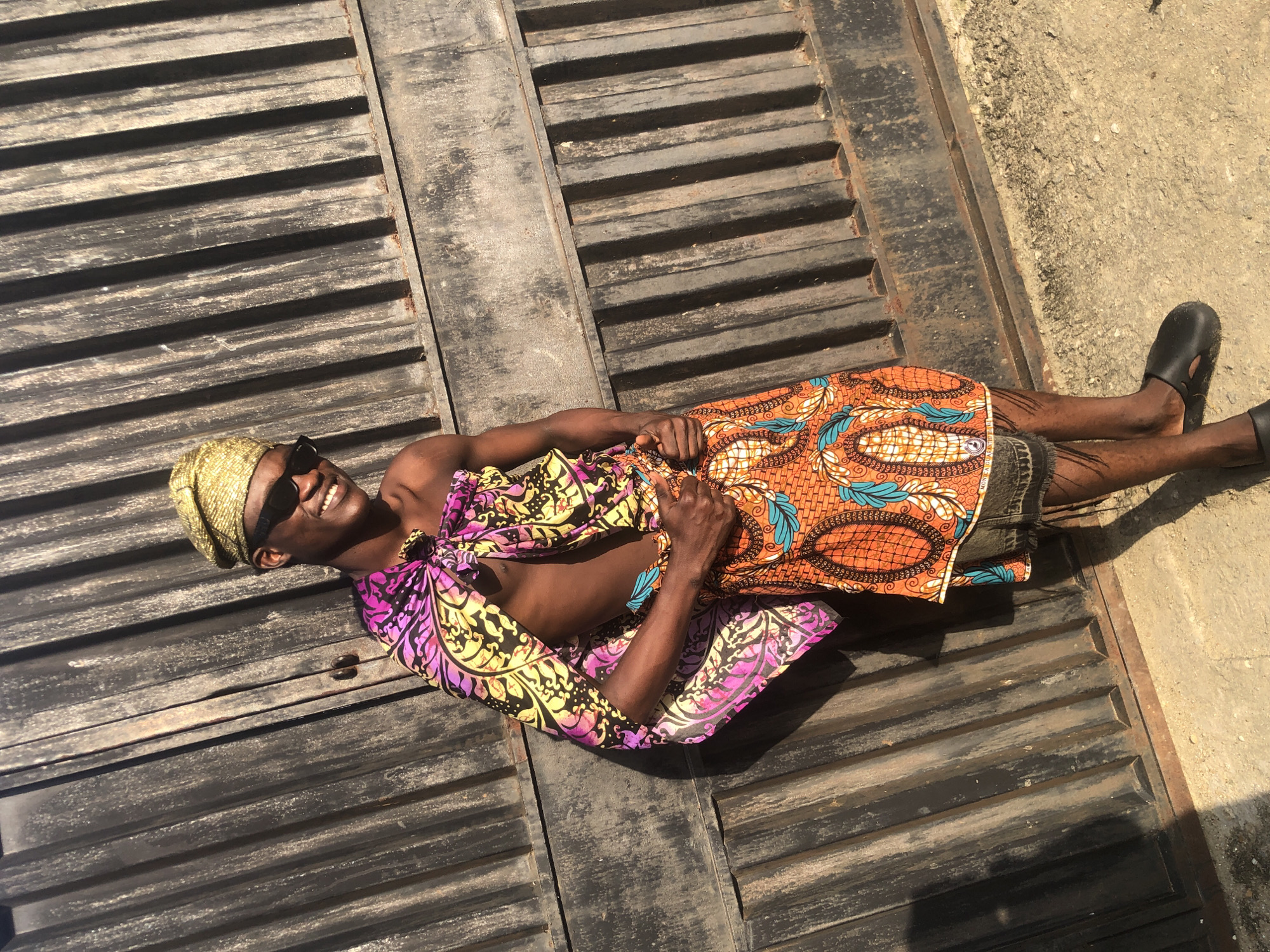 In Ado, Nigeria, a young man stands leaning against a corrugated metal gate. He wears a golden head wrap, dark sunglasses, and a colorful garment draped over his shoulders and chest. The garment features a vibrant pattern of pink, purple, yellow, and black. Below this, he wears a patterned orange skirt with blue and white accents and a fringe at the bottom, revealing denim shorts underneath. He is barefoot, wearing black slip-on shoes. The man smiles at the camera, giving a thumbs-up with his right hand. The background is dominated by the weathered metal gate, with sunlight casting shadows and highlighting the texture of the corrugated surface. The ground beneath him is a rough, sandy concrete.
