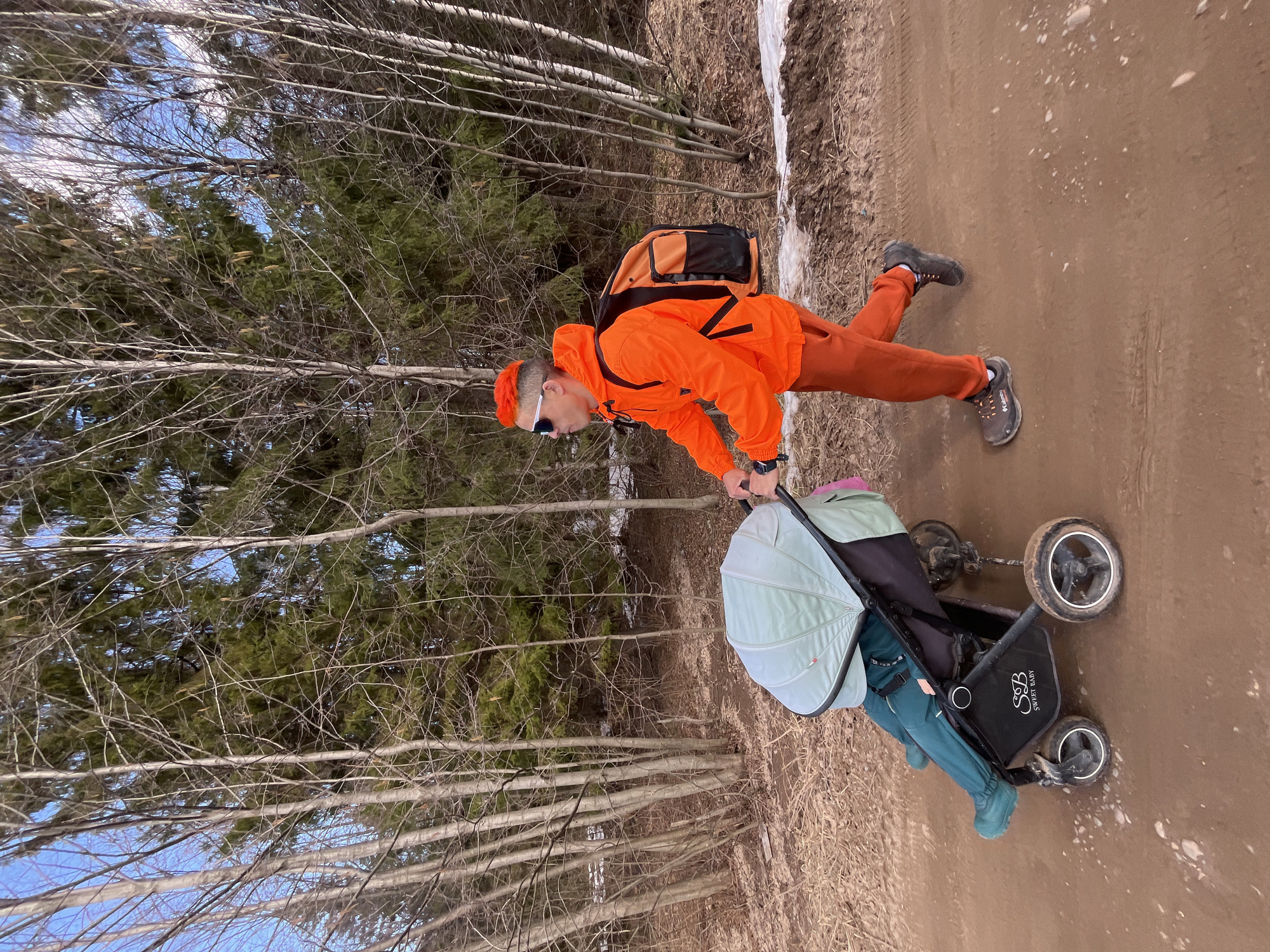 The image depicts a person walking a baby stroller on a dirt path in a wooded area. The person is wearing an orange jacket and pants, sunglasses, and a backpack. The stroller is black with a light green sunshade and appears to be occupied by a baby or toddler. The path is muddy, and there is some snow visible on the side of the path. The trees on either side of the path are bare, with some evergreen trees in the background. Based on research, this image was most likely taken in the city of Chaikovsky, Russia.