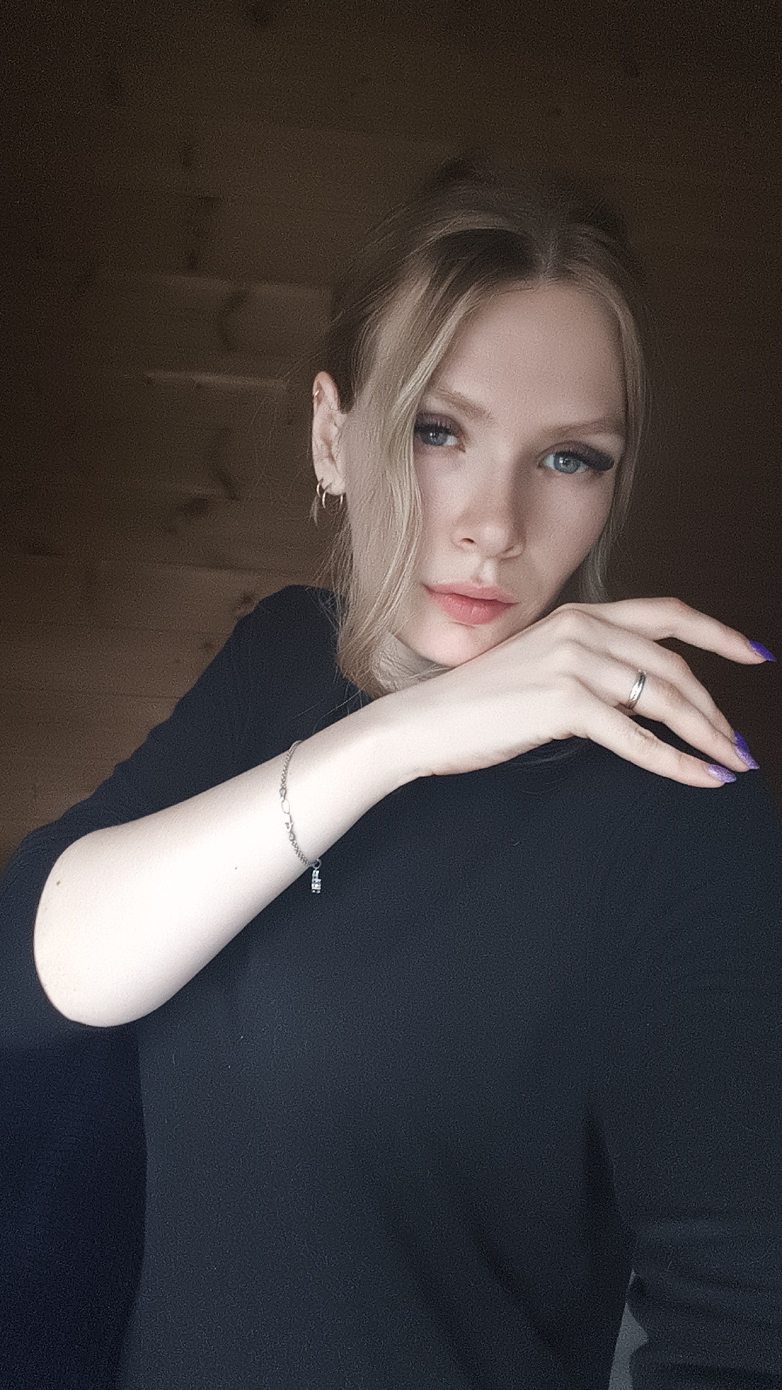 A young woman with blonde hair and blue eyes poses indoors against a dark wooden wall. She wears a black long-sleeved top. Her left arm is visible, adorned with a silver bracelet. Her hand rests near her face, displaying a silver ring and light purple nail polish. She looks directly at the camera with a slightly serious expression. The setting appears to be a dimly lit interior with rustic wooden paneling in the background. The location of Русский Вожой, Russia, cannot be confirmed from the image.