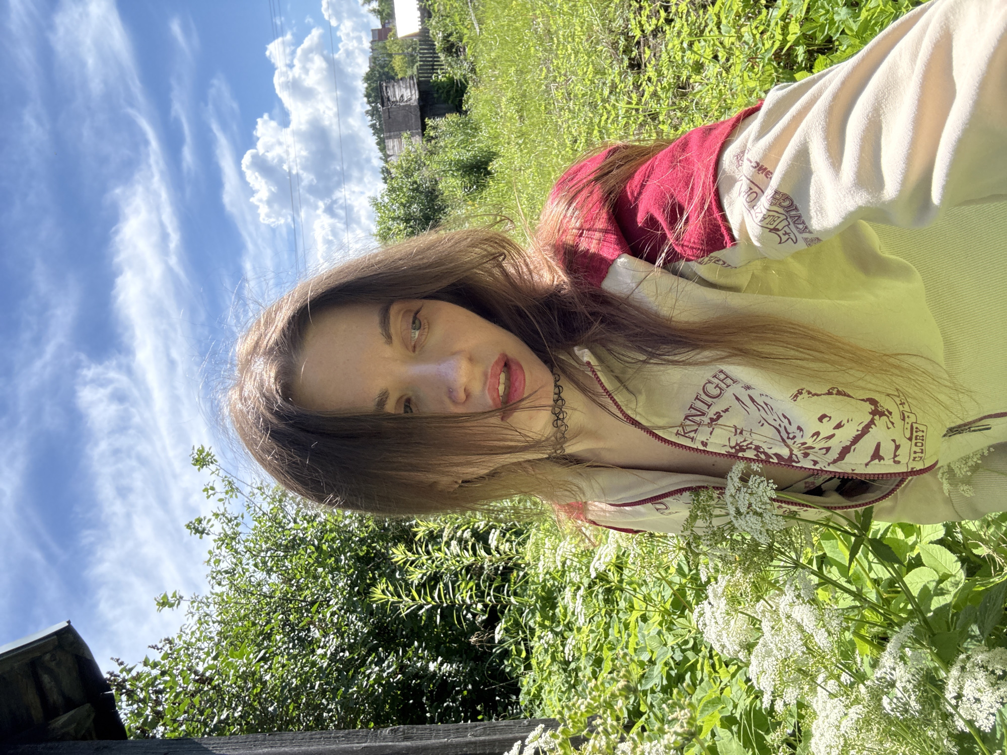 A young woman with long, brown hair and light skin poses for a selfie in a natural outdoor setting. She is wearing a light-colored zip-up jacket with red sleeves and text and graphics on the front. She has a choker necklace on. The background is filled with lush green vegetation, including tall grass, leafy plants, and white flowers. In the distance, there are some rustic wooden structures and a fence, suggesting a rural or semi-rural environment. The sky above is blue with wispy white clouds. The lighting indicates it is daytime, likely sunny, casting natural shadows.