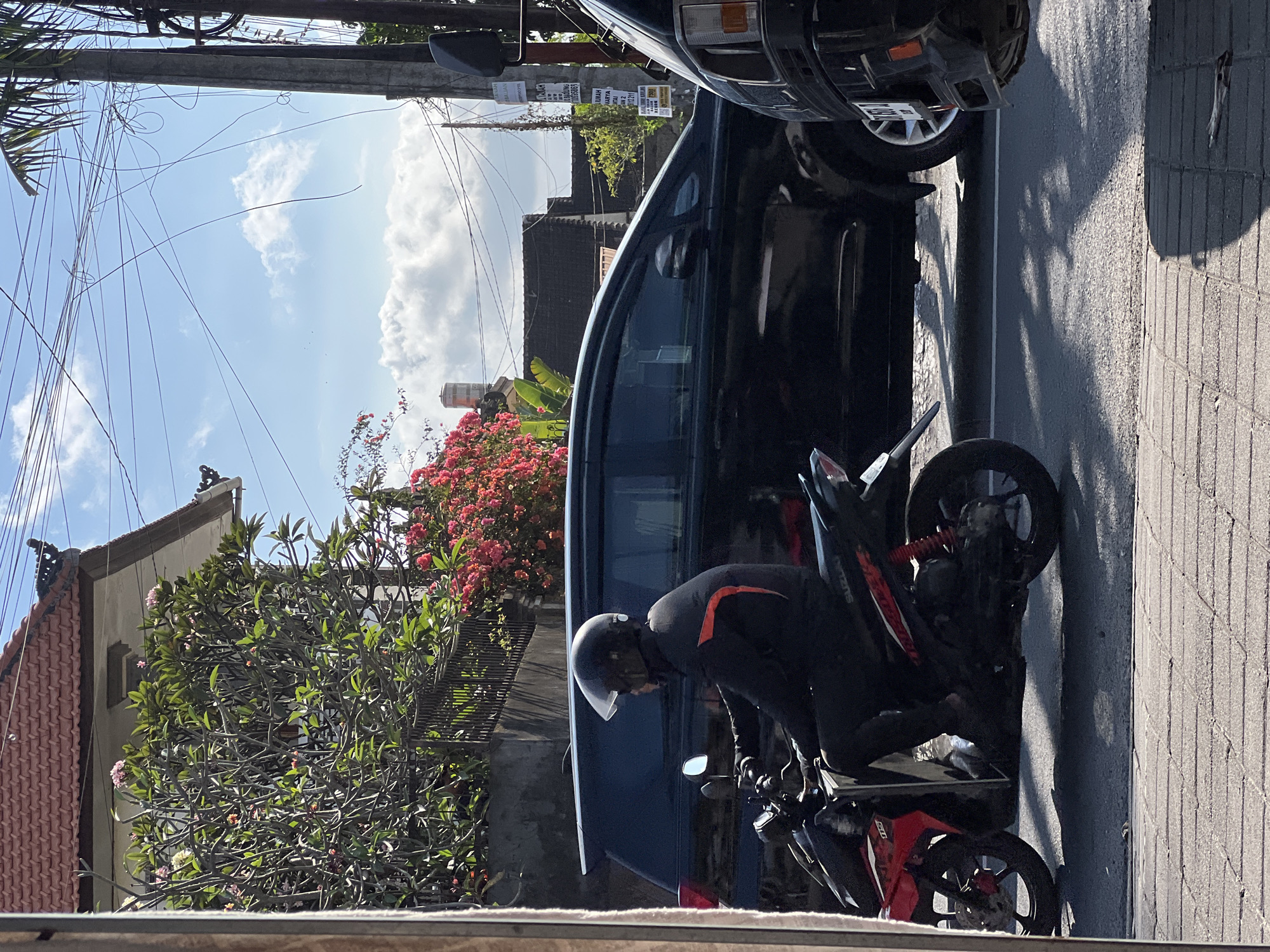 The image depicts a street scene in Denpasar, Indonesia, likely during daylight hours with bright sunlight casting shadows. In the foreground, a person wearing a black jacket, black pants, and a black helmet is riding a red and black motorcycle. The motorcycle has the brand "SUZUKI" visible. The rider's right leg is slightly extended, and they appear to be wearing black shoes. The motorcycle is positioned on an asphalt road, with a tiled sidewalk to the left.
Behind the motorcycle, a dark-colored SUV is parked. The SUV has tinted windows and a roof rack. Further in the background, a building with a red tiled roof and white walls is visible, partially obscured by lush green foliage and vibrant pink flowers. Overhead, a tangle of electrical wires crisscrosses the bright blue sky, which is dotted with scattered white clouds. A utility pole stands to the right of the frame. Signs are affixed to the utility pole, one of which appears to be an advertisement. The overall atmosphere suggests a typical urban environment in Bali.
