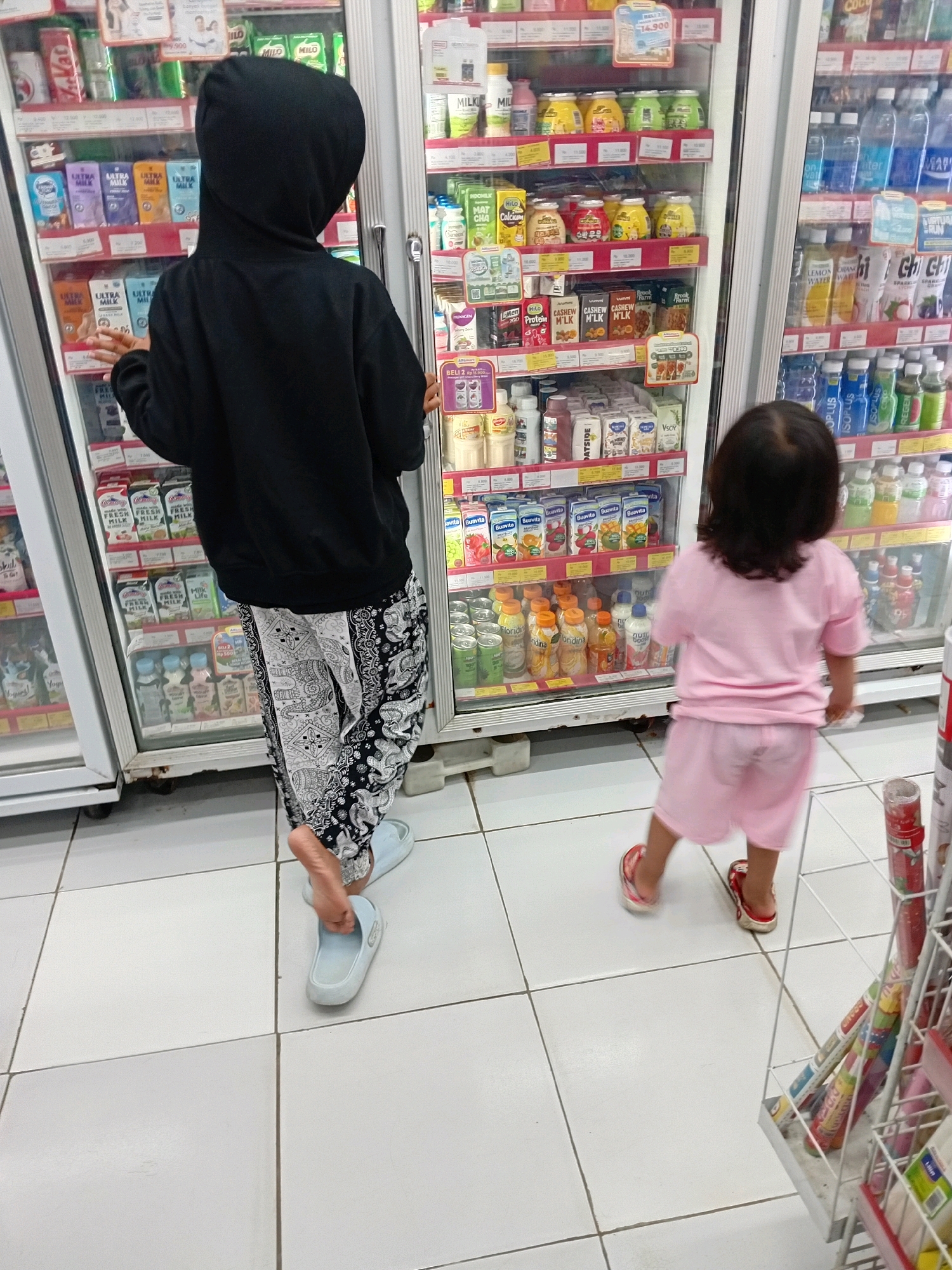 Two individuals, an adult wearing a black hooded sweatshirt and patterned pants, and a child wearing a pink t-shirt and shorts, are standing in front of refrigerators in what appears to be a convenience store. The adult is facing away from the camera, looking into one of the refrigerators. The child is facing away from the camera as well, looking into a different refrigerator to the right of the adult. The refrigerators are filled with various bottled drinks and cartons. The floor is tiled white. The scene is indoors, likely in Seren, Indonesia, based on the provided context.