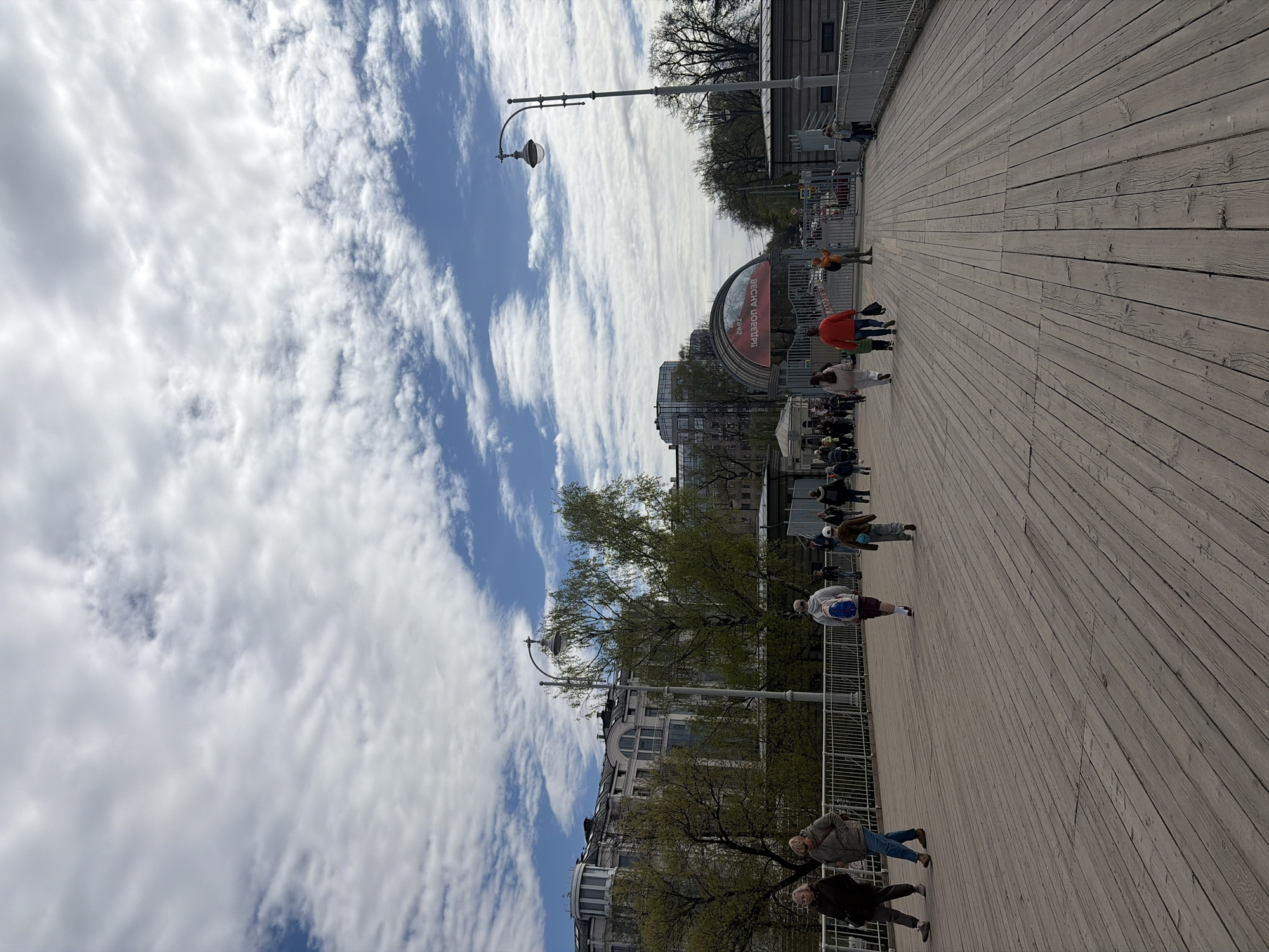 The image shows a view of the wooden boardwalk at New Holland Island in Saint Petersburg, Russia. Several people are walking along the boardwalk. A large archway with lettering (in Russian) can be seen in the background. Trees and buildings line the street in the background. Above the buildings, the sky is visible with patchy clouds. The image is taken from a low angle, making the boardwalk prominent in the foreground. A metal railing runs along the edge of the boardwalk. Streetlights are visible along the walkway.