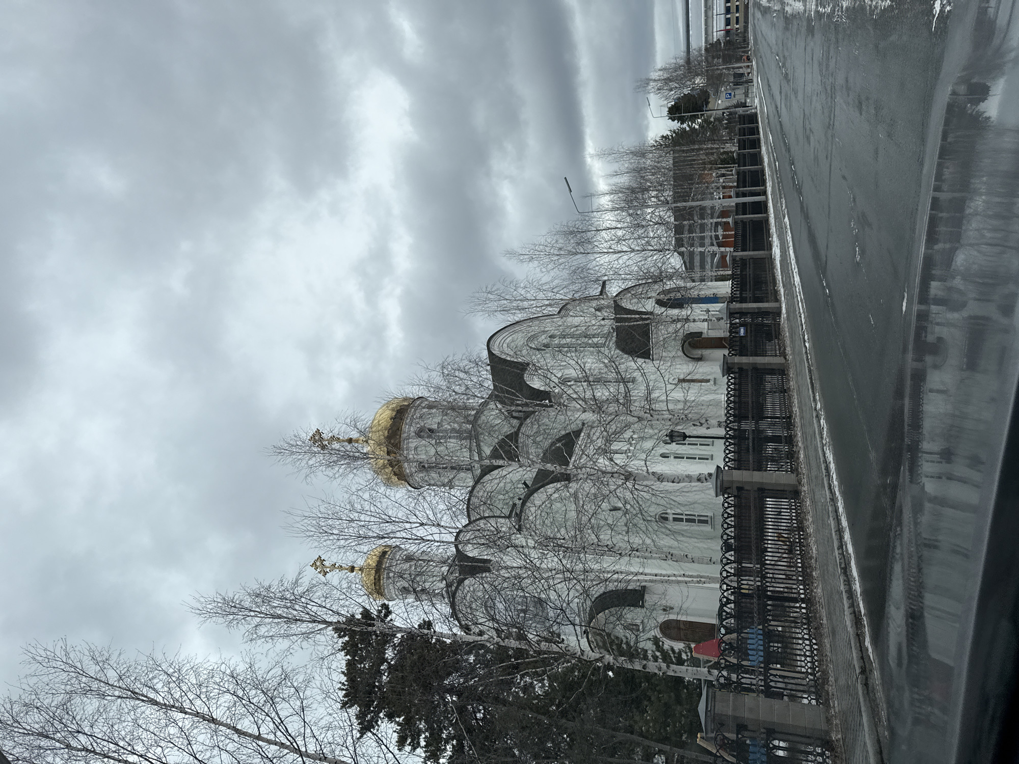 The image depicts a cloudy day in Beloyarsky, Russia. A white Orthodox church with golden domes stands behind a black metal fence. Bare trees obscure parts of the church. A wet road runs alongside the church, reflecting the overcast sky. Buildings are visible in the background.