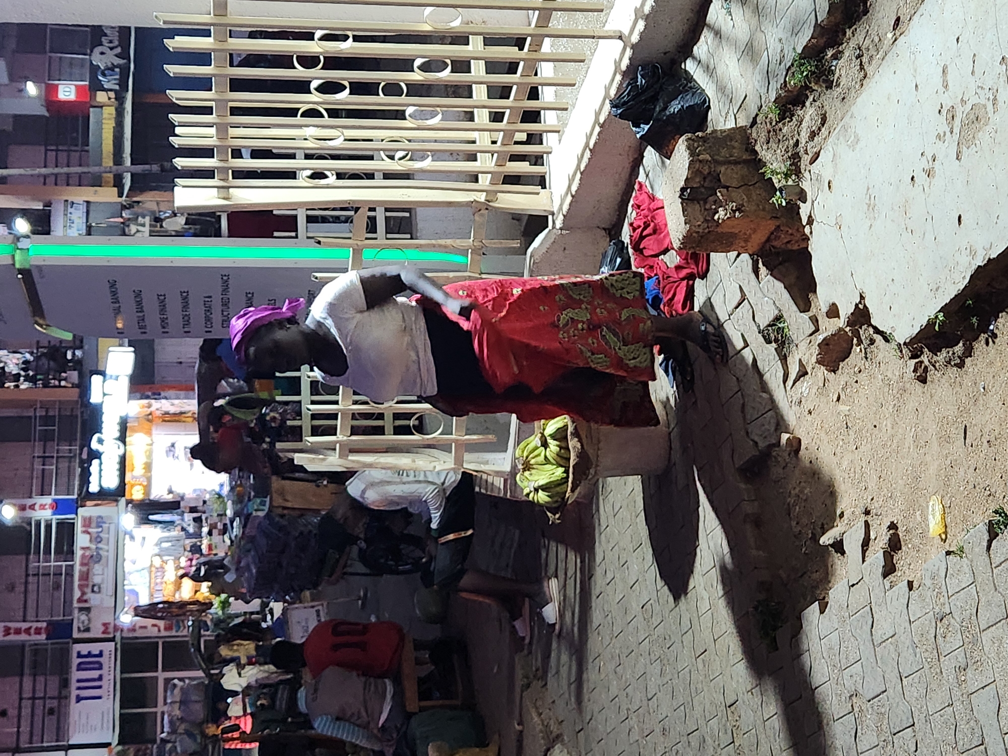 Here is a description of the image:
The image shows a scene on a street in an urban environment, likely a city in an unspecified location. An adult female with a purple headscarf, a white short-sleeved shirt, and a red patterned skirt appears to be engaged in street vending. To her side is a basket filled with green produce, which appears to be green bananas. The woman is positioned near a low concrete barrier that is partially broken or damaged. There is a metal structure behind her, possibly part of a storefront or building. The street is crowded with other individuals and stalls, indicating a bustling commercial area. The signs "TILDE" and "WEAR" are visible on a building in the background. Several other businesses are lined up, with bright light and a sign that reads "RETAIL BANKING," "TRADE FINANCE," "CORPORATE," and "STRUCTURED FINANCE" indicating a financial institution is nearby. The pavement is patterned with cobblestones. The image appears to be taken at night, with artificial light sources illuminating the scene.