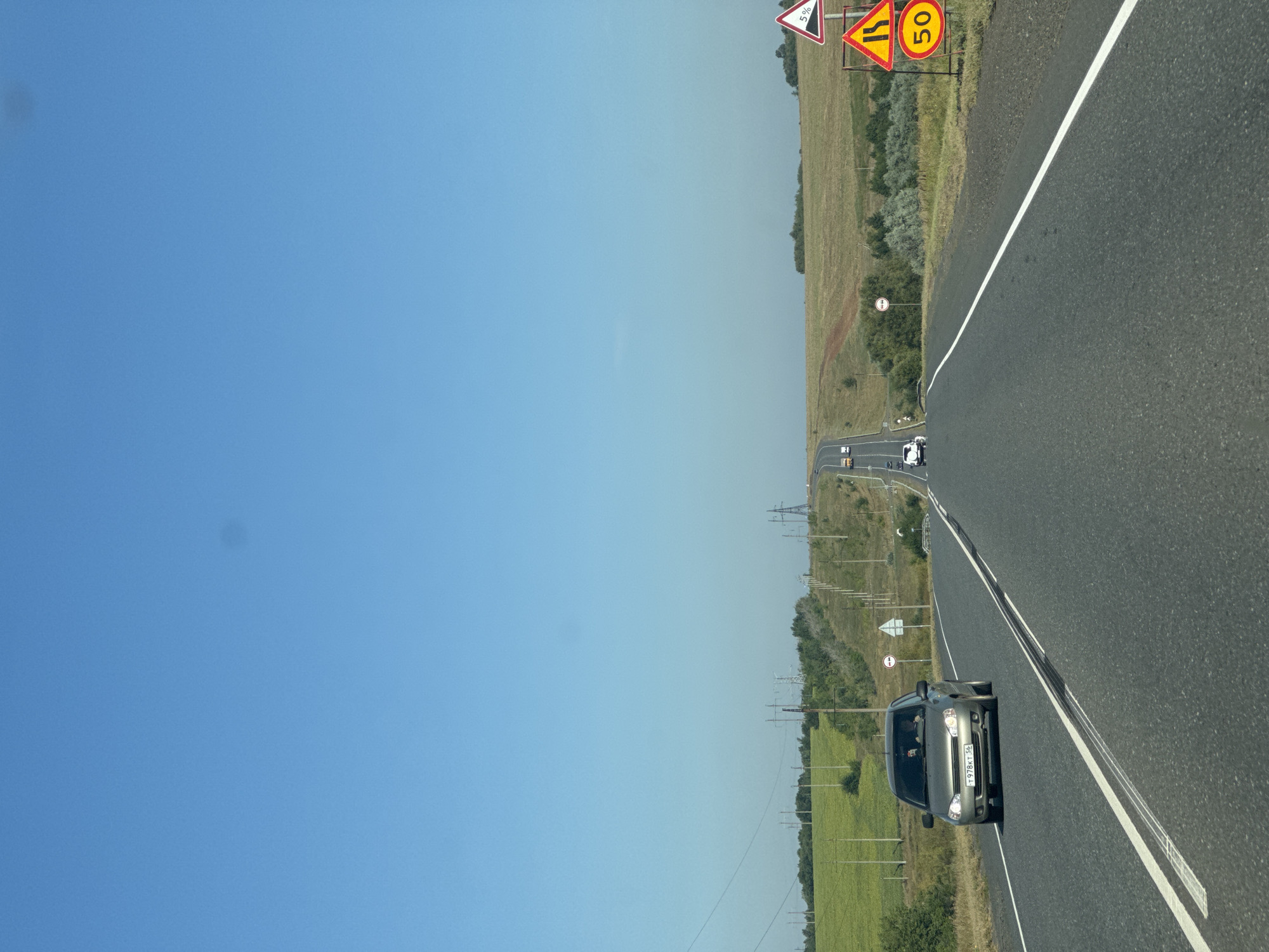 The image captures a view from a vehicle traveling on a paved road heading towards a distant horizon under a clear blue sky. The road itself is marked with double white lines, indicating two lanes of traffic. On the right side of the road, several traffic signs are visible, including a sign with "5%" indicating a steep descent and another circular yellow sign with "50" signifying a speed limit.
The landscape on either side of the road is characterized by rolling hills covered in green vegetation, possibly fields. Electrical poles and power lines stretch across the background, indicating infrastructure in the rural setting. A few vehicles are present on the road, with one grey car in the foreground on the left lane, its license plate reading "T 978KT 56". Other cars are seen further down the road in the distance. The overall scene depicts a typical rural highway in Russia on a bright, sunny day. The location is identified as Krasnaya Niva, Russia.