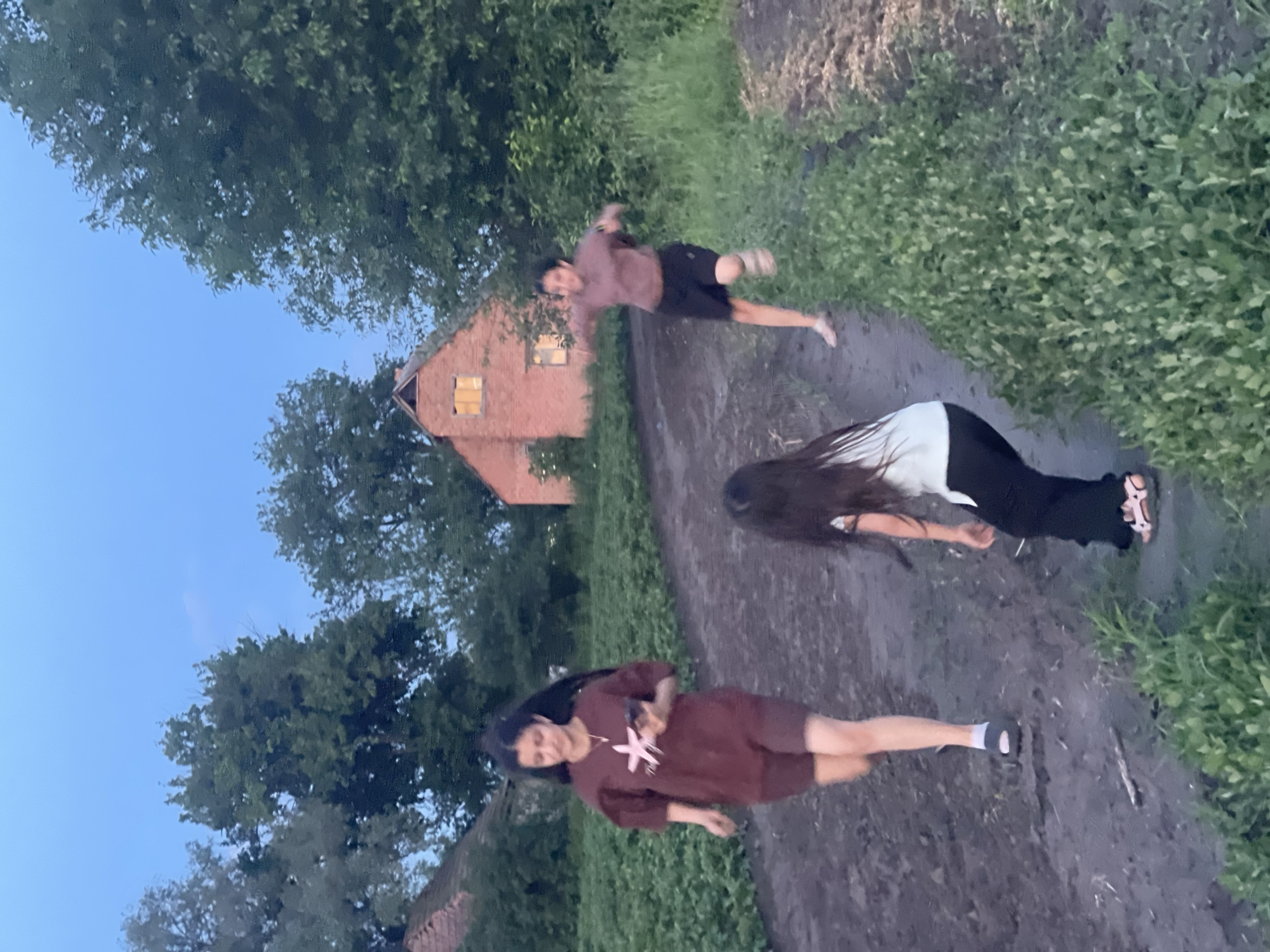 The image captures an outdoor scene in the city of Unknown, Unknown, during dusk or early evening. Three individuals, appearing to be female, are situated on a dirt path flanked by dense green vegetation. A young woman with long dark hair, positioned in the left mid-ground, wears a maroon t-shirt with a light-colored star graphic and matching shorts, dark sandals, and light socks, while holding a smartphone in her right hand and walking forward. In the right mid-ground, another young woman with dark hair wears a light brown top and dark shorts, depicted in mid-stride, running with outstretched arms. A child with long dark hair, visible in the bottom-center foreground, wears a white top, dark pants, and pink sandals, and is shown in a crouched or bending position. In the background, a reddish-brown brick house with a gabled roof and two illuminated windows is prominent. Another structure with a reddish roof is partially visible on the far left. The sky is a clear light blue. The path exhibits an uneven, possibly damp, texture, with rows of low-lying green plants on the left and taller, wilder vegetation on the right.