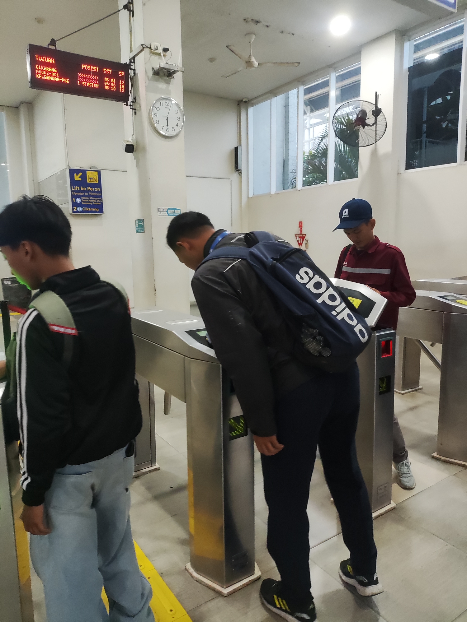 The image depicts an indoor scene at what appears to be a train station in Telagamurni, Indonesia. In the foreground, two individuals are seen passing through automated turnstiles. The person on the left, viewed from the back, wears a black jacket with white stripes on the sleeves and light blue jeans. They are carrying a backpack. The person in the center, also viewed from the back, is wearing a dark jacket and dark pants, and is also carrying a backpack with "adidas" branding. This individual is leaning over a turnstile, interacting with its screen. To the right, a third person wearing a maroon shirt and a dark blue cap is standing at another turnstile, looking down at it.
In the background, a digital display board shows train departure information with destinations like "CIKARANG," "ANGKE-JRT," and "KP.BANDAN-PSE." A clock with a white face and black numbers is mounted on the wall to the left of the digital board. There are also signs indicating "Lift ke Peron" (Elevator to Platform) with platform numbers 1 and 2. The environment appears to be a clean, modern station with tiled floors and white walls. Fluorescent lighting illuminates the space, and a ceiling fan and a wall-mounted fan are visible. The overall impression is of a functional transit hub.