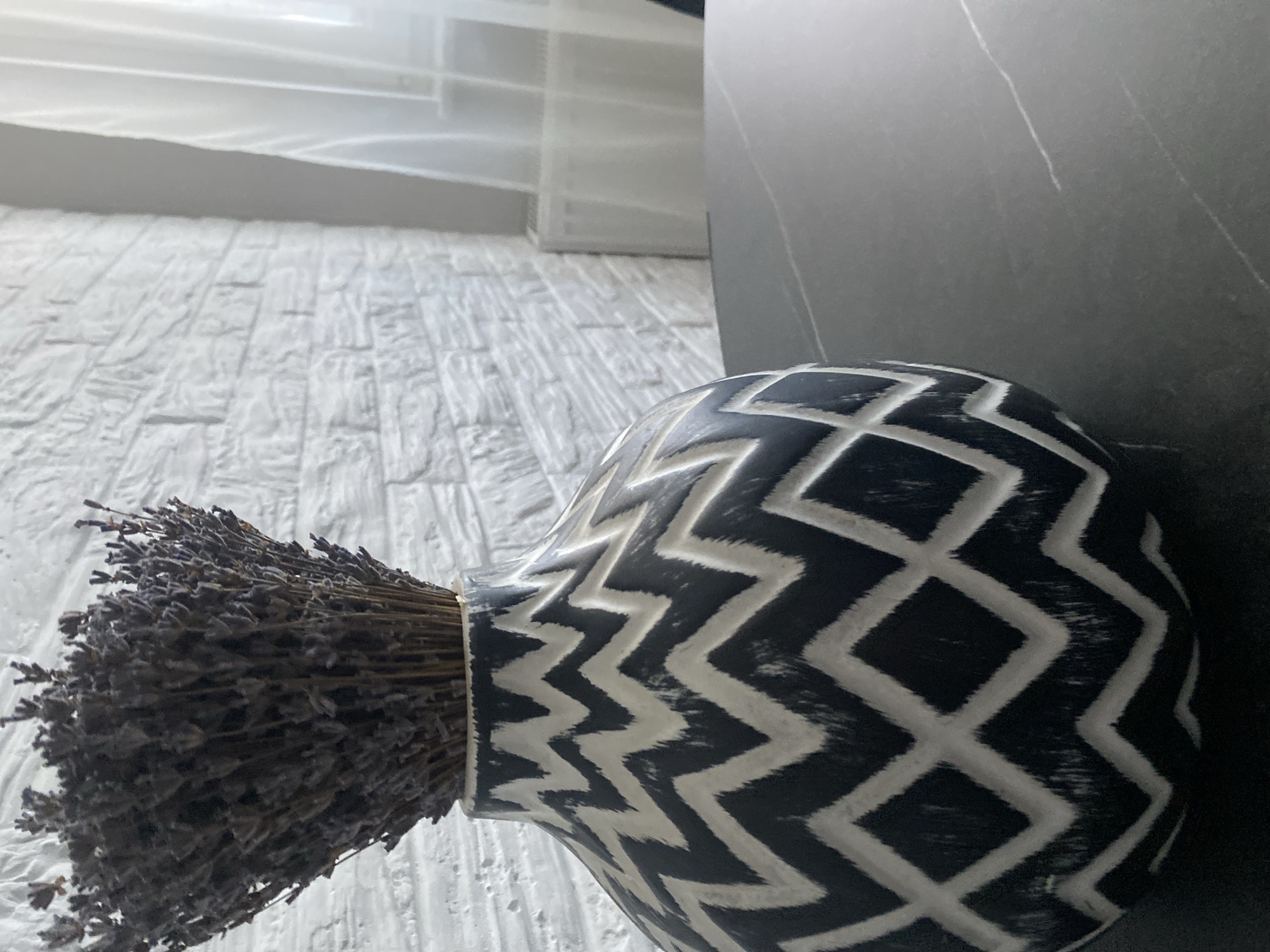 An indoor scene shows a close-up of a patterned vase on a dark, speckled surface. The vase is black with a white, jagged geometric pattern. It is filled with dried lavender. In the background, a white brick wall is visible, with a sheer white curtain and a radiator to the right. The lighting suggests it is daytime, possibly from a window. There are no people or animals present. The specific location in Chișinău, Moldova is not discernible from the image.