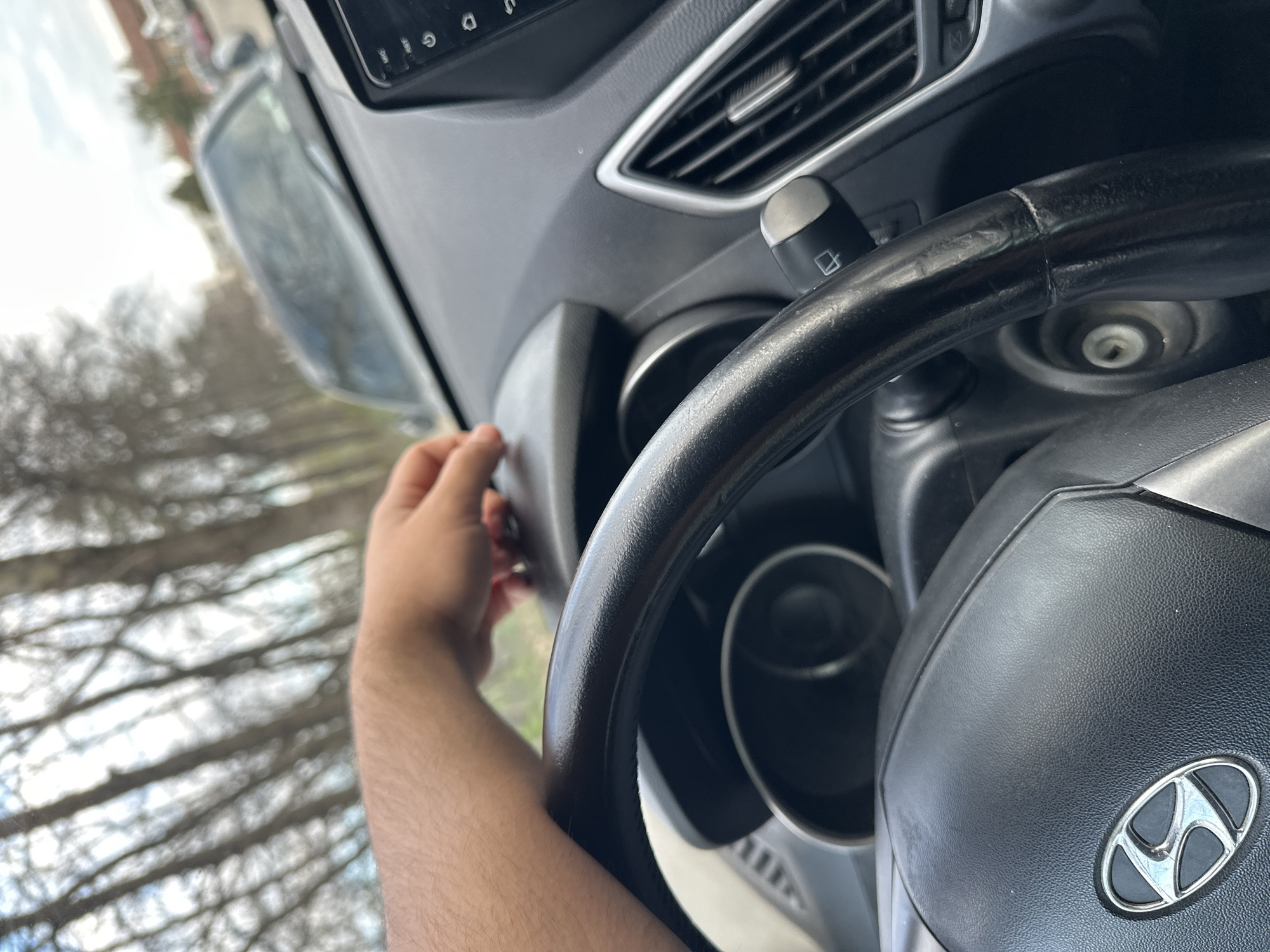 A person's hand is reaching towards the dashboard of a car, possibly inserting a key or adjusting a control. The steering wheel is visible with a Hyundai logo. The car's interior also includes air vents and a turn signal stalk. The exterior shows trees and possibly a building, suggesting the car is either parked or moving in an area with foliage. No specifics about the location within Plovdiv, Bulgaria, can be determined.