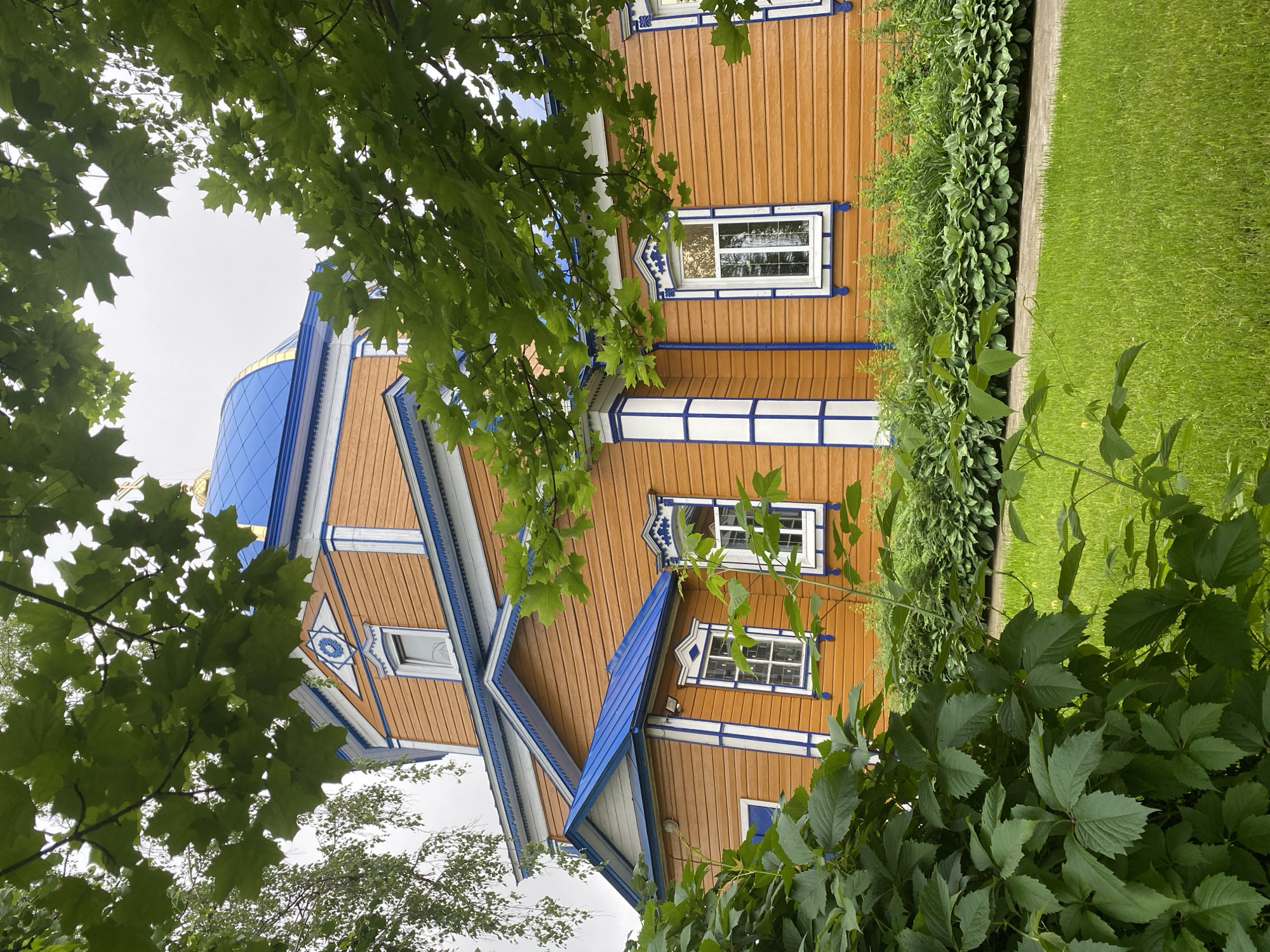 The image shows a bright orange wooden church with a blue domed roof, framed by lush green foliage and trees. The church has white trim with blue accents around the windows and along the rooflines. A well-maintained lawn and garden beds with various plants surround the building. The sky is overcast, indicating a cloudy day. The scene is peaceful and serene, with the natural elements complementing the architectural beauty of the church. The church is located in the city of Zelenodol'sk, Russia.