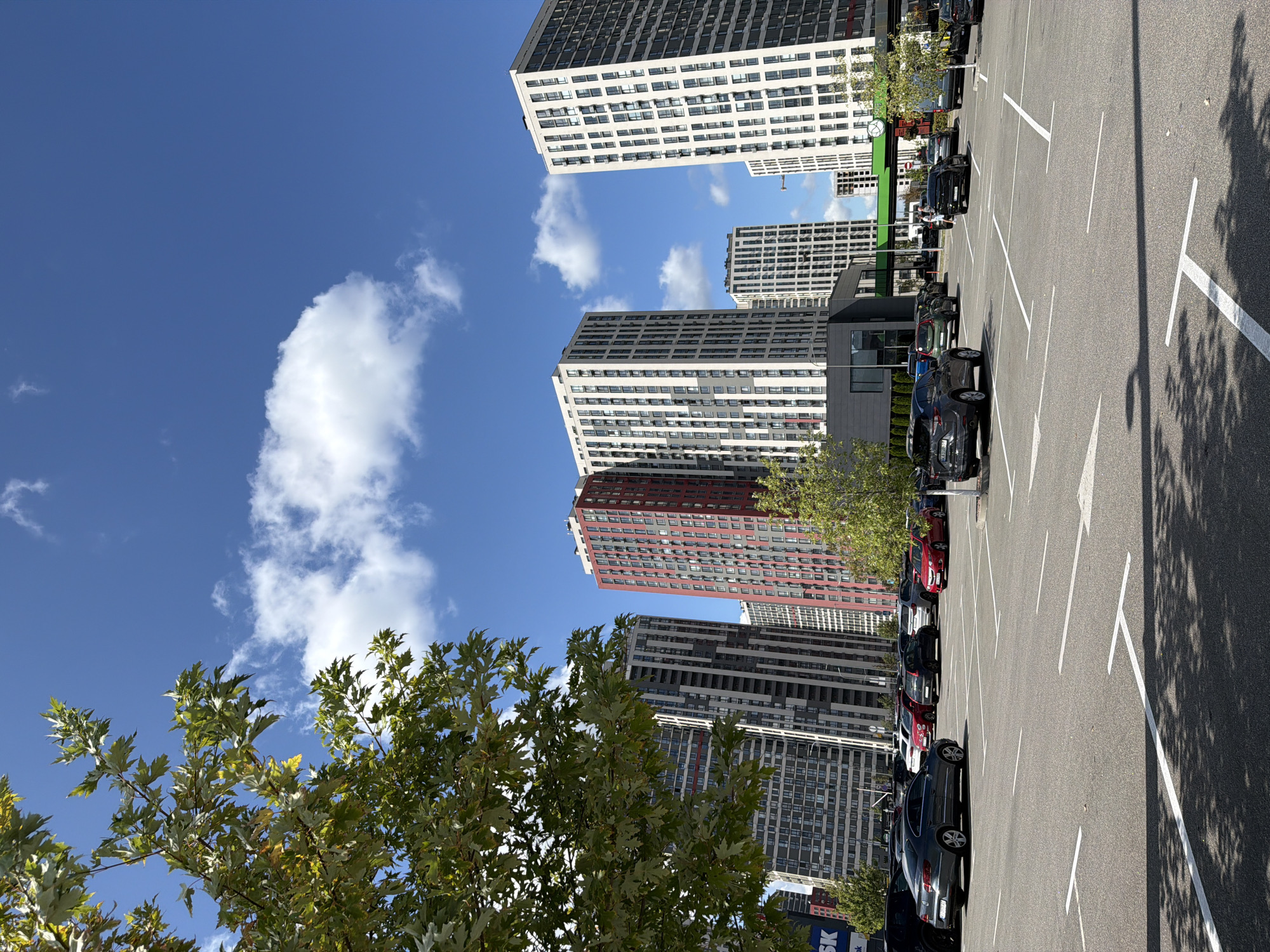 A wide-angle, eye-level shot shows a parking lot in front of several modern, high-rise residential buildings, likely in Kyiv, Ukraine, on a clear day. A large tree with green leaves partially obstructs the view on the left side of the frame. The buildings are tall and feature diverse architectural styles, with some displaying red and gray facades and others a predominantly white and gray color scheme with numerous windows. The parking lot is asphalted and marked with white lines indicating parking spaces and directional arrows. Several cars are parked in the lot, including a dark gray sedan in the foreground on the left, a red sedan on the right, and other vehicles of various colors scattered throughout. The sky is bright blue with scattered white clouds. The overall impression is of a modern urban environment.