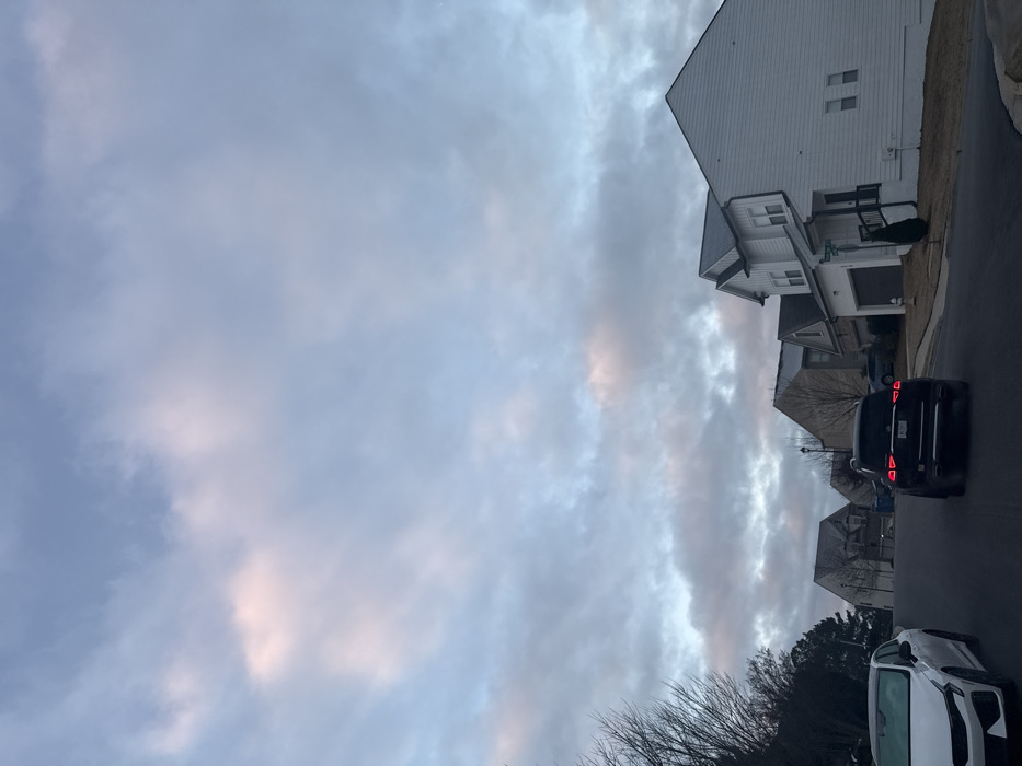 This image depicts a suburban residential street scene at what appears to be dawn or dusk in Lawrenceville, Georgia, United States. The location is identifiable by visible street signs reading "SUMMIT DR" and "WESTFIELD TRCE" at the intersection on the right.
The sky is largely overcast with stratocumulus clouds, exhibiting tones of gray, blue, and faint pink/peach, indicative of low sun angle. The environment consists of a gently sloping asphalt road leading uphill. On both sides of the street are multiple two-story detached single-family houses, characteristic of modern suburban architecture, featuring various siding colors and dark roofs.
In the foreground on the left, a white Kia K5 sedan is parked facing away from the viewer, exhibiting what appears to be a light layer of frost or dew on its surfaces. In the center of the road, a dark-colored SUV, possibly a Lexus or Volkswagen, is positioned with its brake lights illuminated, suggesting it is either stopped or in the process of moving forward. Further down the street on the right, a prominent white house with dark trim and a dark roof stands at the aforementioned intersection. A fire hydrant is visible on the curb near this house, along with a small evergreen shrub. Beyond this, another vehicle is partially visible parked in a driveway. A dark-colored trash or recycling bin is visible on the left side of the road, past the moving SUV. Trees lining the street and properties are bare, indicating a winter or early spring season, and the ground cover is dormant grass. No individuals are visible in the scene.