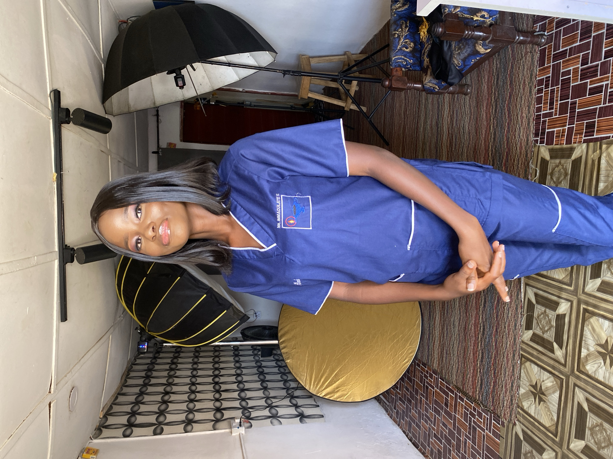A young woman is captured in a full-length portrait, standing indoors, likely in Vom, Nigeria. She is wearing a navy blue scrub uniform with white trim on the collar, sleeves, and pants, suggesting a medical or healthcare profession. Her dark hair is styled long and straight with a grey streak on one side. She poses with her hands clasped loosely in front of her, looking directly at the camera with a slight smile and light makeup.
The setting appears to be an indoor space that doubles as a makeshift photo studio. Various photography lighting equipment is visible, including two large black diffusion umbrellas – one a standard umbrella and another a hexagonal softbox – and a round gold reflector disc resting on the floor.
The background features an eclectic mix of surfaces. To the left, there's a light-colored wall with vertical panels. To the right, the wall has patterned brown and white tiles. The floor is covered with two different types of flooring: a patterned dark rug or carpet and a section of brown and white tiled floor with a geometric star-like pattern. A wooden chair with blue and gold upholstered cushions is partially visible in the upper right background.
On the left chest of her uniform, embroidered text is visible, reading "MR. IMMACULATE R." with a logo featuring a blue shape and a small red circle. The overall impression is a professional portrait taken in a somewhat informal or adaptable studio environment.