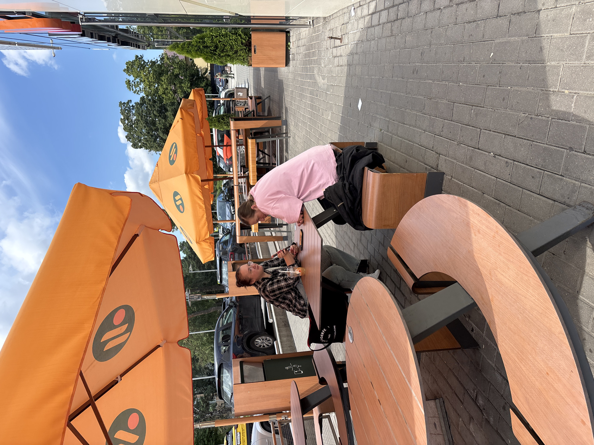 Two young women are seated at an outdoor table under a large orange umbrella. The woman on the left is wearing a plaid shirt and jeans and is looking towards the right while holding a drink. The woman on the right is wearing a pink t-shirt and is looking down at the table. The table is made of wood and has a dark gray base. They are sitting on a bench that is also made of wood and has a dark gray base. The area is paved with gray bricks. In the background, there are more outdoor tables and umbrellas, as well as cars and trees. The scene appears to be a cafe or restaurant with outdoor seating. The location is specified as Gorki-2, Russia.