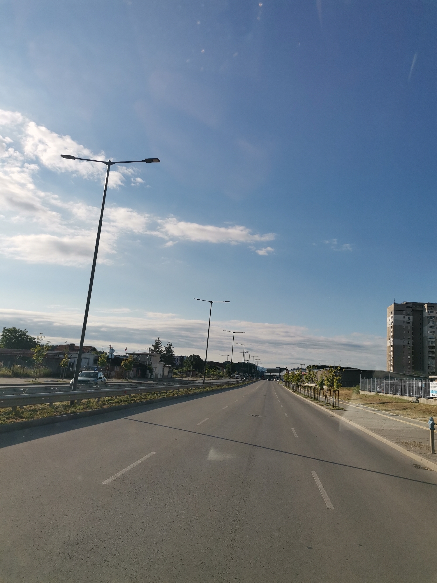 The image shows a straight, empty road in the city of Sofia, Bulgaria, with light traffic. The road is asphalt and has lane markings. There are streetlights on both sides of the road. On the left side, there are some buildings, trees, and a parked car. On the right side, there is a tall, modern apartment building and a sidewalk. The sky is blue with some white clouds. The overall impression is of a clear, sunny day.