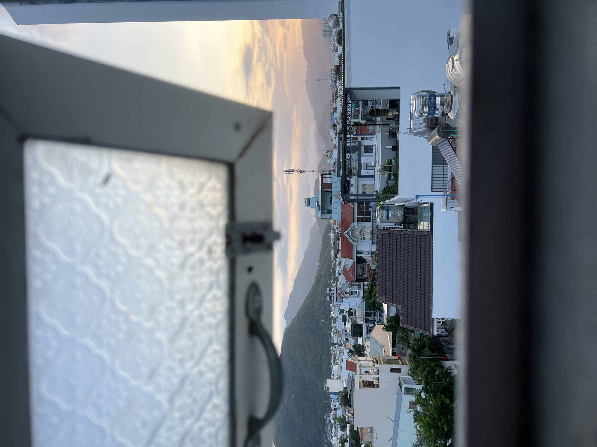 The image depicts a view from a building in Nam Nha Trang Ward, Vietnam, overlooking a densely populated urban landscape situated along the coast, with mountains in the distance. The image is framed by a window, with the window pane visible on the left. Various residential buildings with different roof styles and heights are visible, including some with rooftop water tanks. The sky shows a mix of clouds and sunlight, indicative of either dawn or dusk. A communication tower is discernible amidst the buildings. No people are visible in the frame.