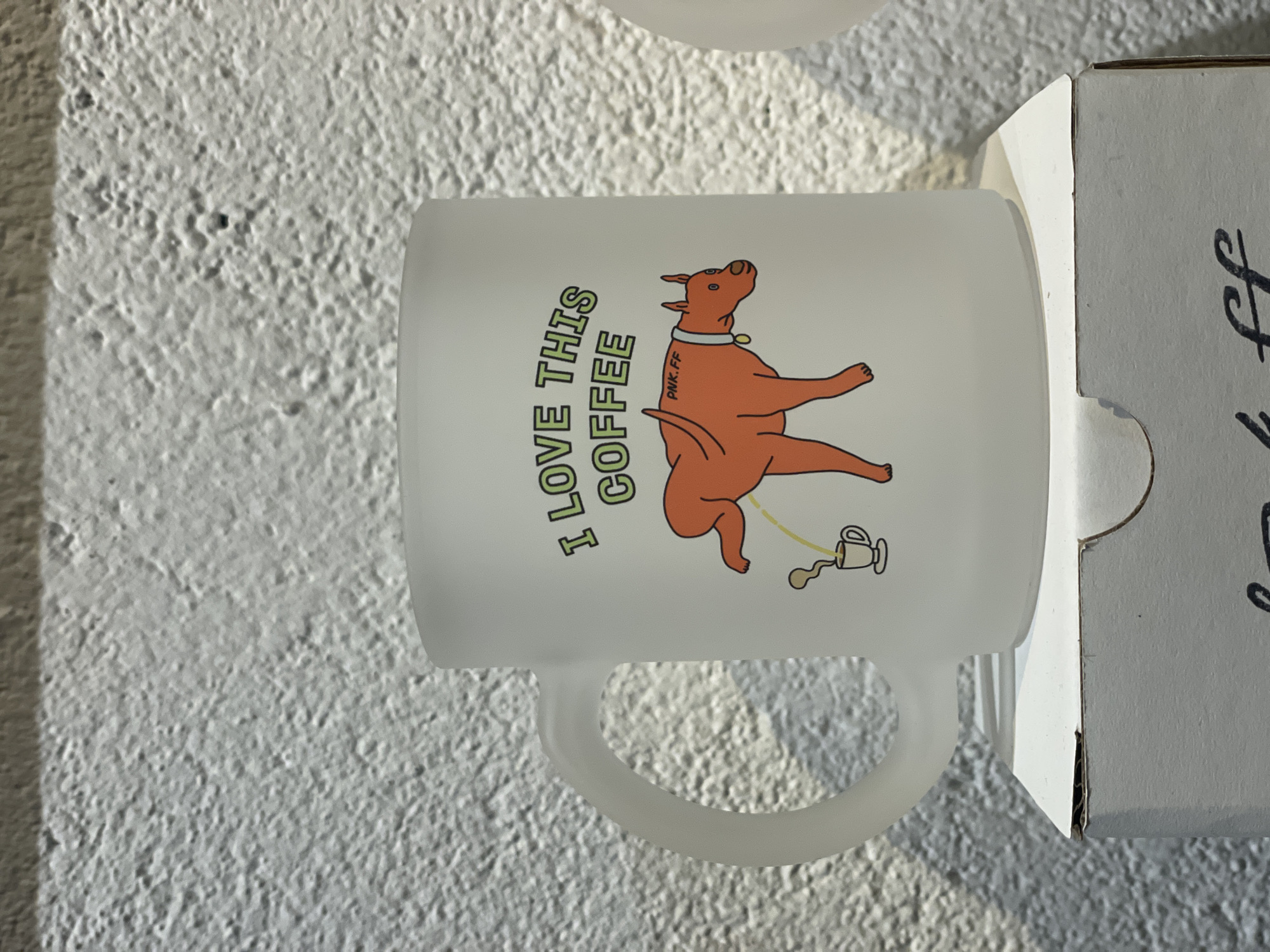 A white mug with a cartoon illustration sits next to its cardboard box on a textured white surface. The illustration shows an orange dog with a collar urinating into a coffee mug, with the words "I LOVE THIS COFFEE" printed vertically on the mug. The mug appears to be resting against the box, with a portion of black text visible on the box's side. The textured white surface could be a wall or a tabletop. The scene does not show specific elements indicating a location in Bangkok, Thailand.