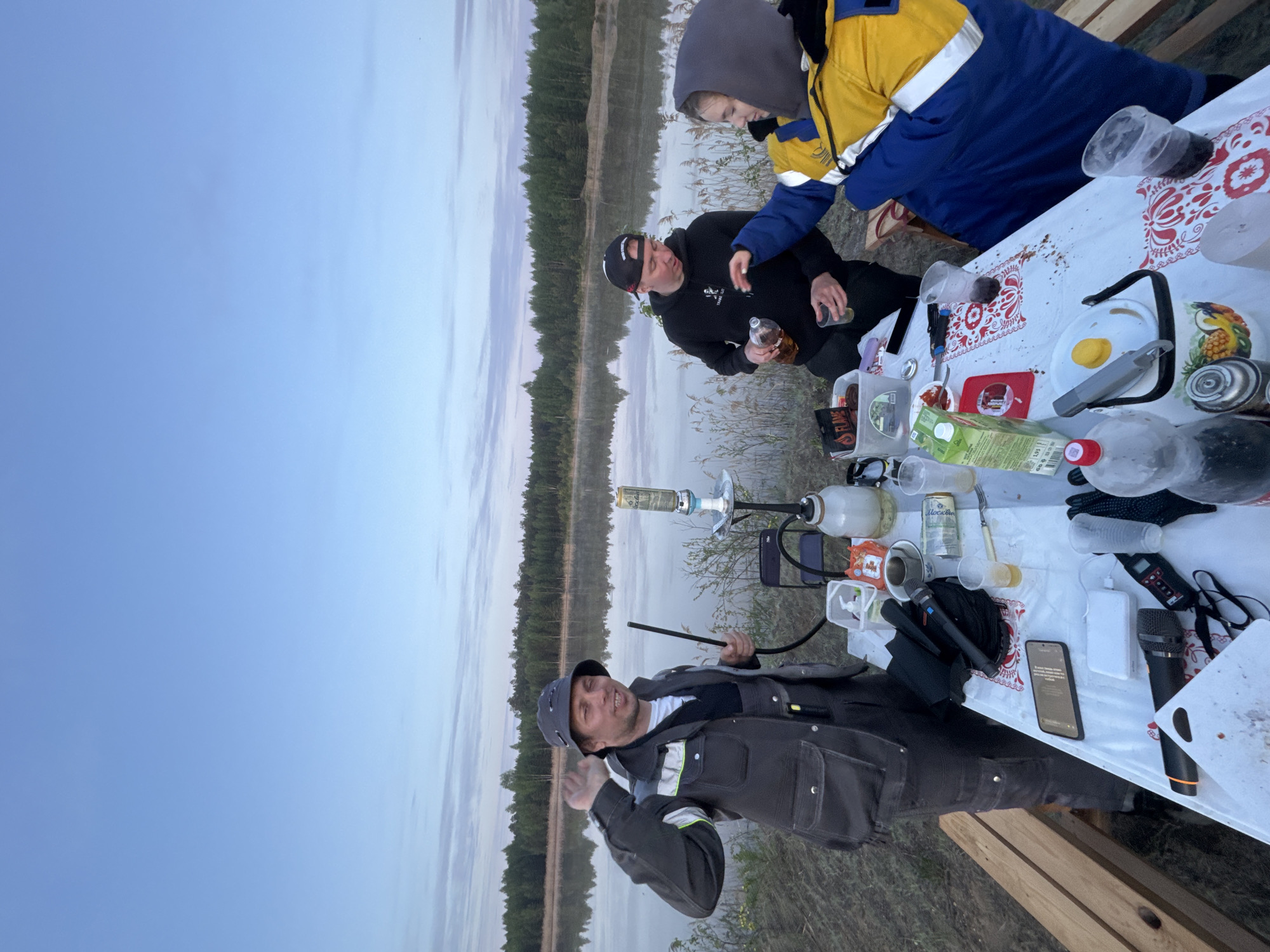 Three people are seated around a white table with a patterned tablecloth at a lakeside gathering in Novyi, Russia. One person wears a dark jacket and hat and holds a hookah hose. Another person, in a black sweatshirt and hat, sits near a person in a blue and yellow jacket. The table is laden with assorted items, including bottles, cups, a juice box, and electronic devices. A calm lake reflecting the sky is visible in the background, with trees lining the opposite shore. The sky suggests twilight or dawn.