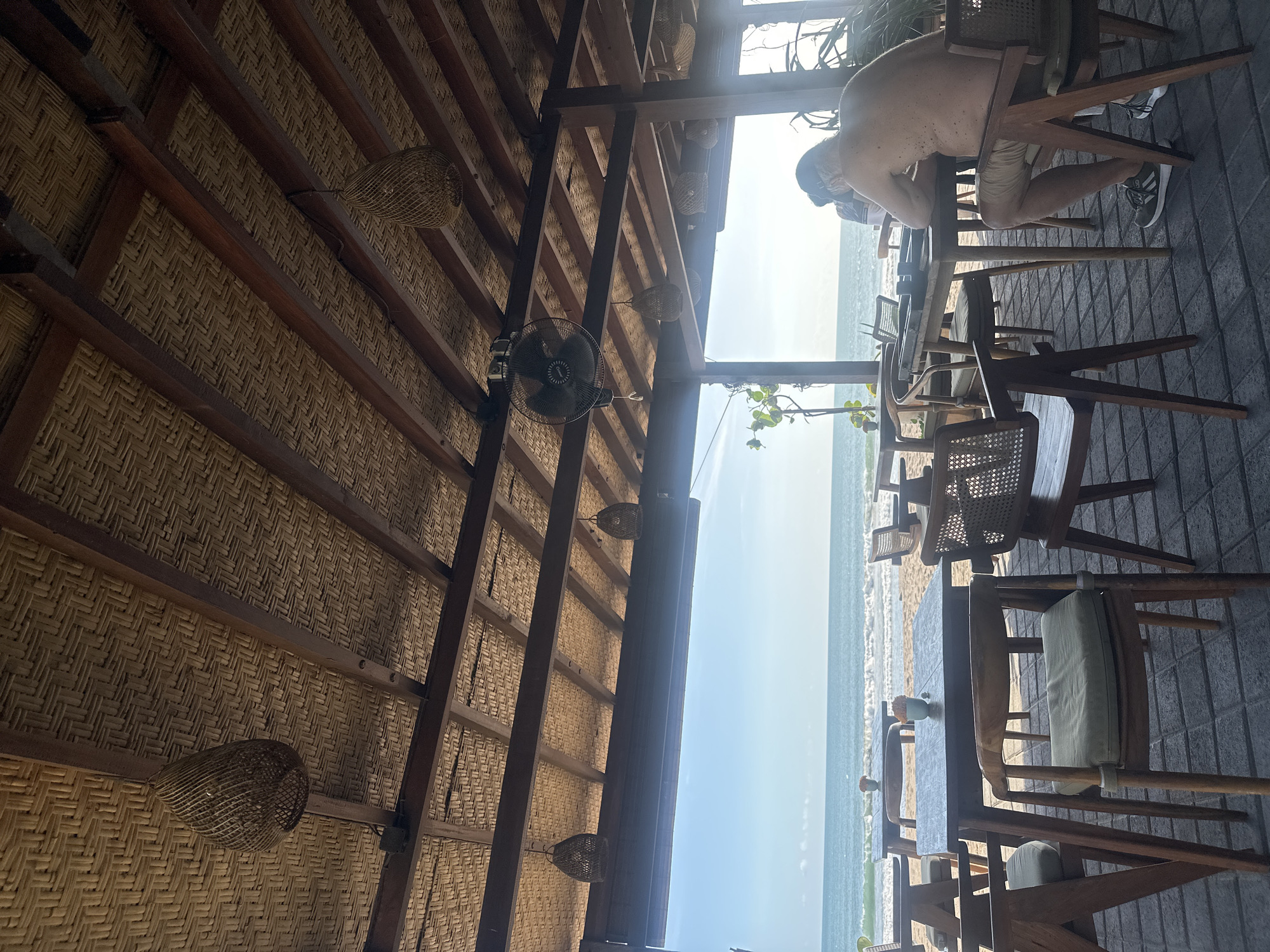 The image shows an outdoor seating area, likely a restaurant or cafe, in North Kuta, Indonesia. The area is under a thatched roof with wooden beams and decorative woven lamps. A black fan is mounted on one of the beams. Outside the seating area, a sandy beach meets a light blue ocean under a bright, clear sky. Tables and wooden chairs are arranged in the foreground, with one table occupied by a man leaning on his arms. He is wearing a face mask, shorts and shoes. The ground is paved with gray tiles.