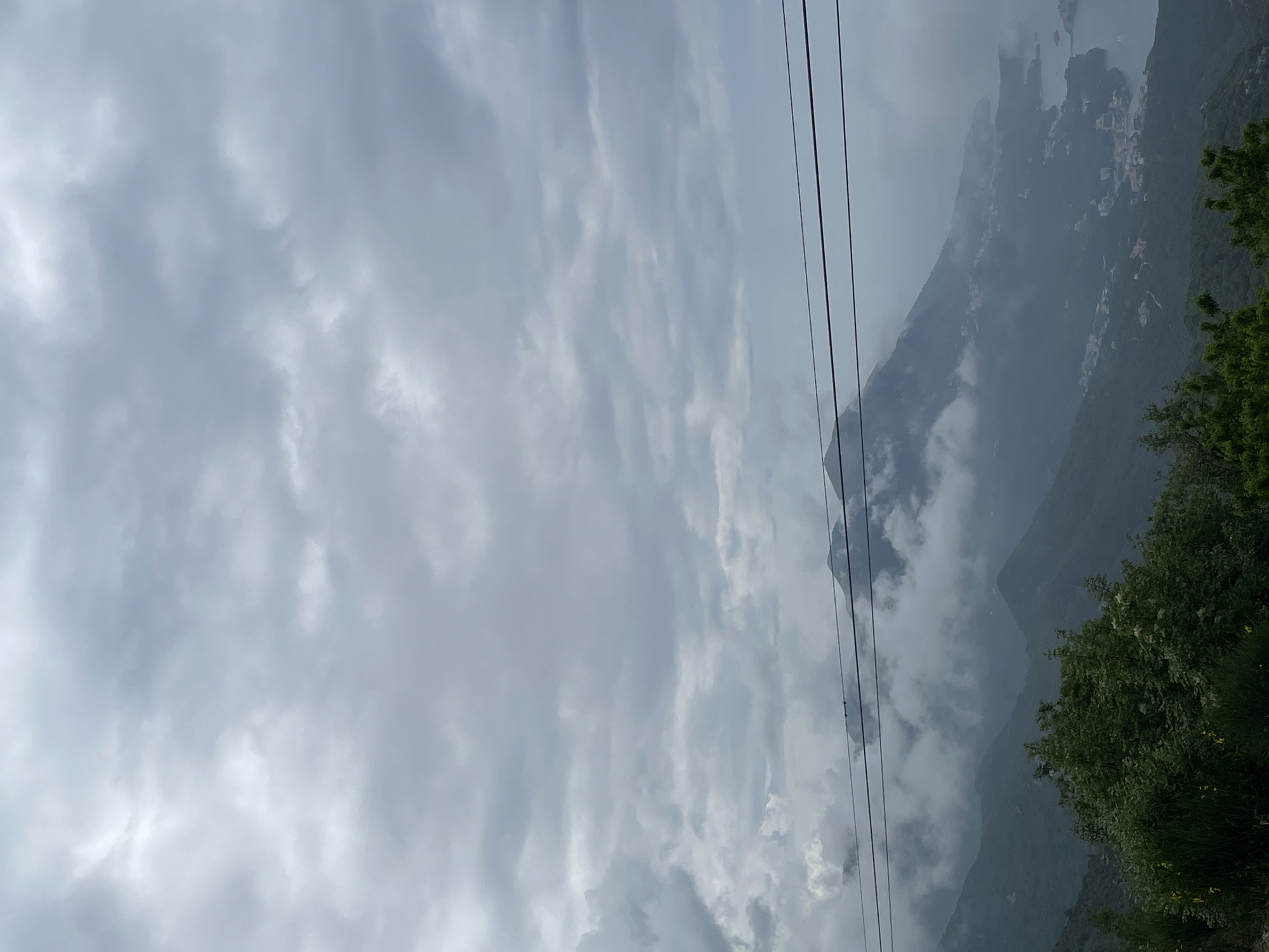 The image depicts a landscape in Brajici, Montenegro, under a heavy, overcast sky filled with grey clouds. Mountains shrouded in mist and fog are visible. Several power lines run diagonally across the view. Vegetation, including trees and shrubs, are situated on the right side of the frame. Buildings can be made out amid the mountainside.