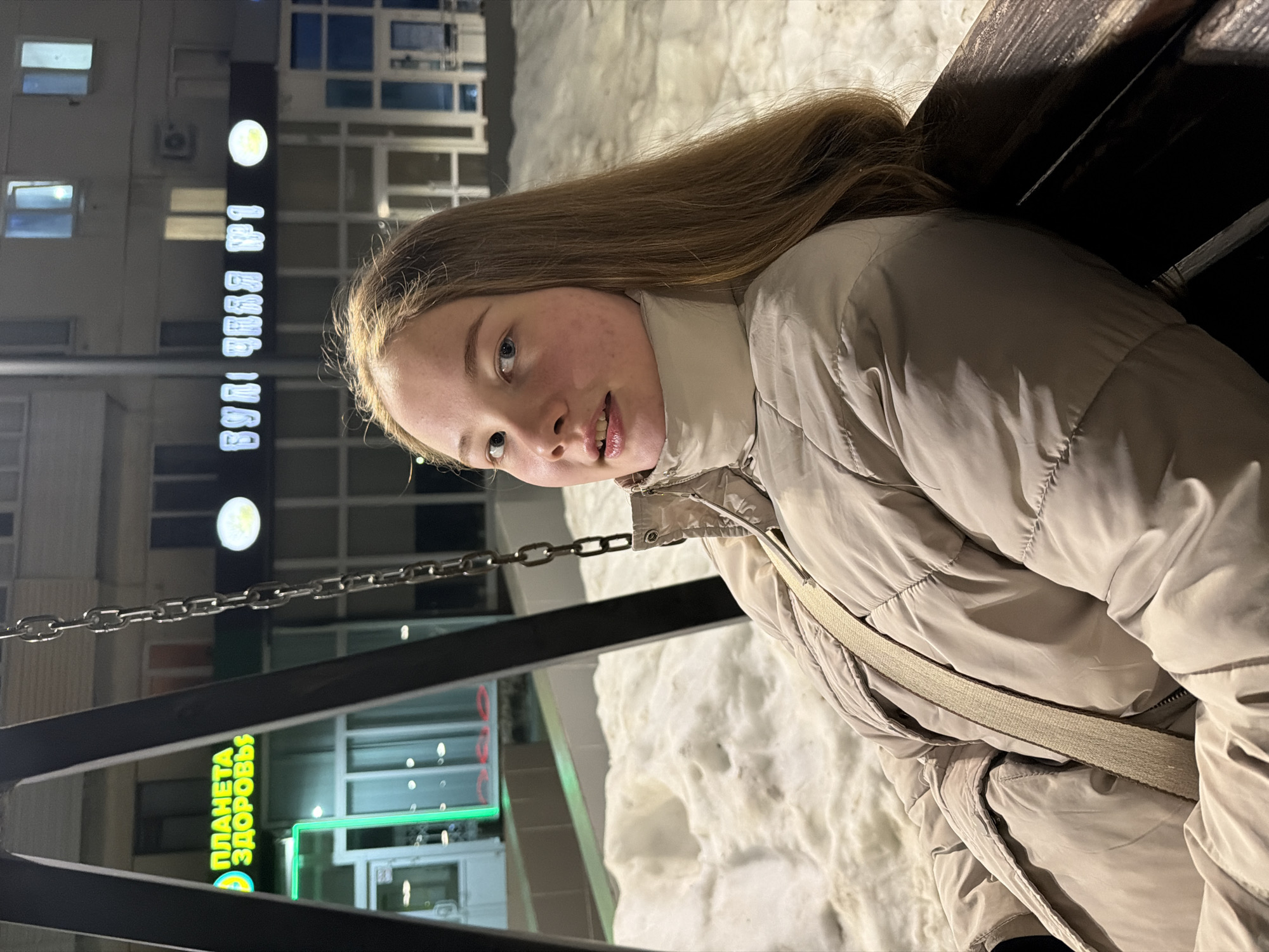 A young woman sits on a swing bench in Chaikovsky, Russia. She wears a beige puffer jacket and has long, brown hair. The background includes a building with a sign reading "PLANETA ZDOROVYYA" (Planet of Health) in green letters, which confirms the location. There is also snow on the ground and a building in the background with lights on.