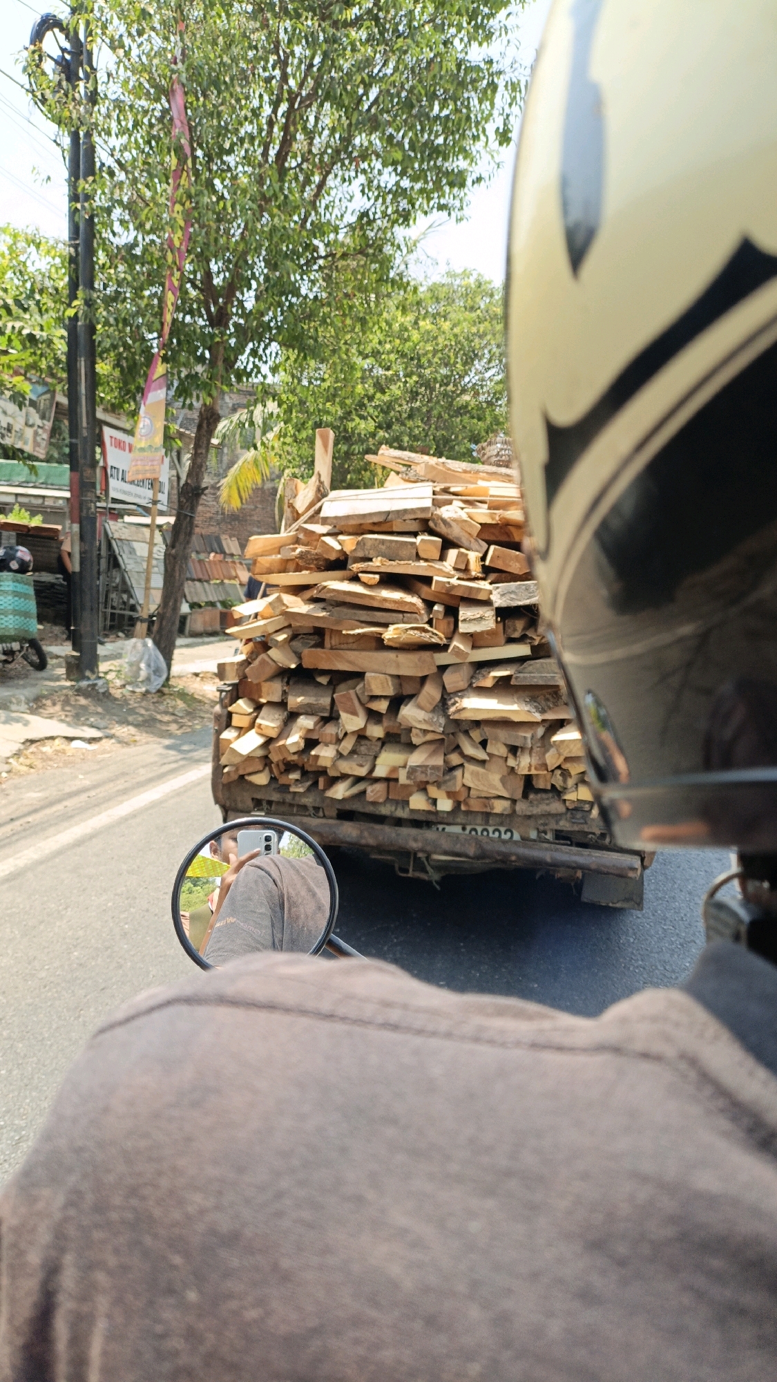 This is a first-person perspective image taken from a motorcycle on a street in Kuwasen, Indonesia. The motorcycle is moving, and the image captures the road ahead, the rear of a truck loaded with wood, and a reflection in the motorcycle's side mirror.
The foreground is dominated by the back of the rider's dark brown shirt and the rider's helmet, which is partially visible on the right side of the frame. The helmet is white and black.
In the middle ground, a truck is seen from behind, heavily laden with a large pile of cut lumber. The wood appears to be of various sizes and shapes, stacked haphazardly. The truck itself looks utilitarian, with a visible license plate that is partially obscured but appears to contain numbers.
To the left of the truck, a sidewalk and roadside environment are visible. There is a utility pole with wires, trees with green foliage, and some buildings or structures in the background. One building has a sign with text that is difficult to read clearly but appears to be in Indonesian. There is also a small green cart or basket on the left side.
The reflection in the motorcycle's side mirror provides a clearer view of the rider, who is wearing a brown shirt, and a glimpse of the environment to the side, including some greenery and potentially a building or stall. The reflection shows the rider's face, though it is somewhat indistinct.
The overall scene depicts a typical streetscape in a built-up area, with traffic and commercial activity implied by the presence of the truck and buildings. The lighting suggests it is daytime, with bright sunlight casting shadows.