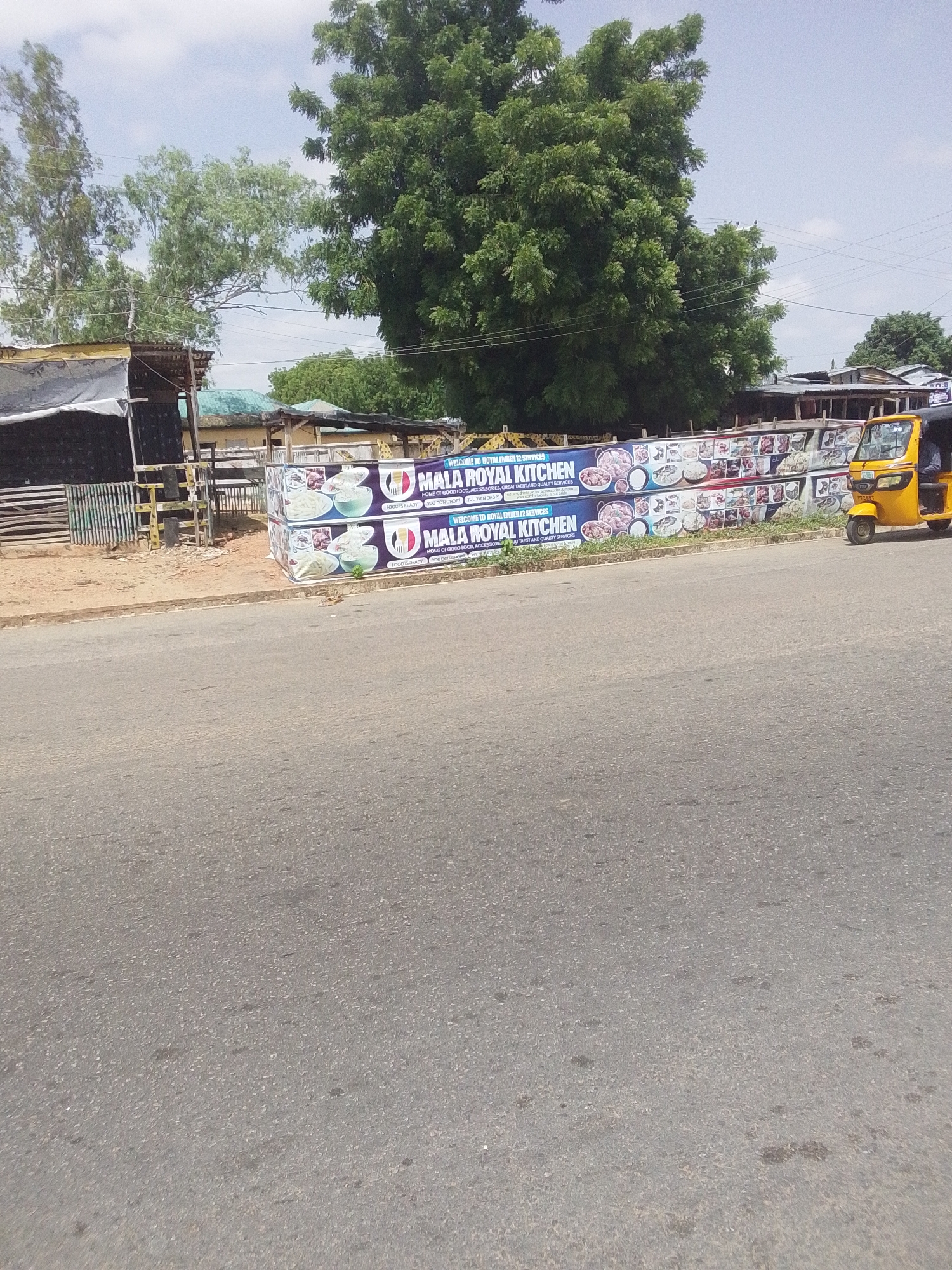 The image shows a street scene in Jalingo, Nigeria, featuring a prominent banner for "MALA ROYAL KITCHEN." The banner, stacked two high, displays images of various dishes and advertises "GOOD FOOD, ACCESSORIES, GREAT TASTE AND QUARTZ SERVICES." To the right of the banner, a yellow auto-rickshaw, a common mode of transport in Nigeria, is parked. The background consists of trees, some overhead wires, and various structures, suggesting a roadside establishment. The road itself is paved and appears to be a typical street in a Nigerian city. The sky is partly cloudy.