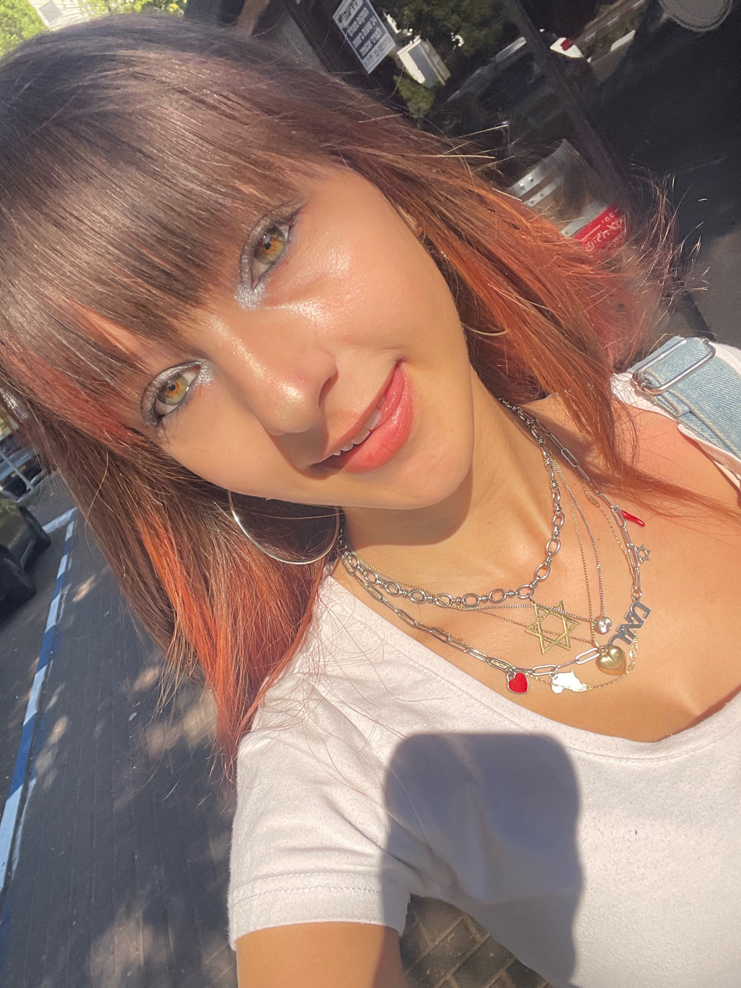 A young woman with auburn hair and bangs, her eyes are golden-green and have metallic silver eyeshadow. She wears a white t-shirt and denim overalls. She is adorned with several necklaces, one with a Star of David, a golden heart, and a red chili pepper charm. Her mouth is slightly open, revealing her teeth, and she appears to be smiling. The background shows a street scene in Raanana, Israel, with shadows of trees and what appears to be a building. The lighting suggests it is daytime, possibly late afternoon due to the long shadows.