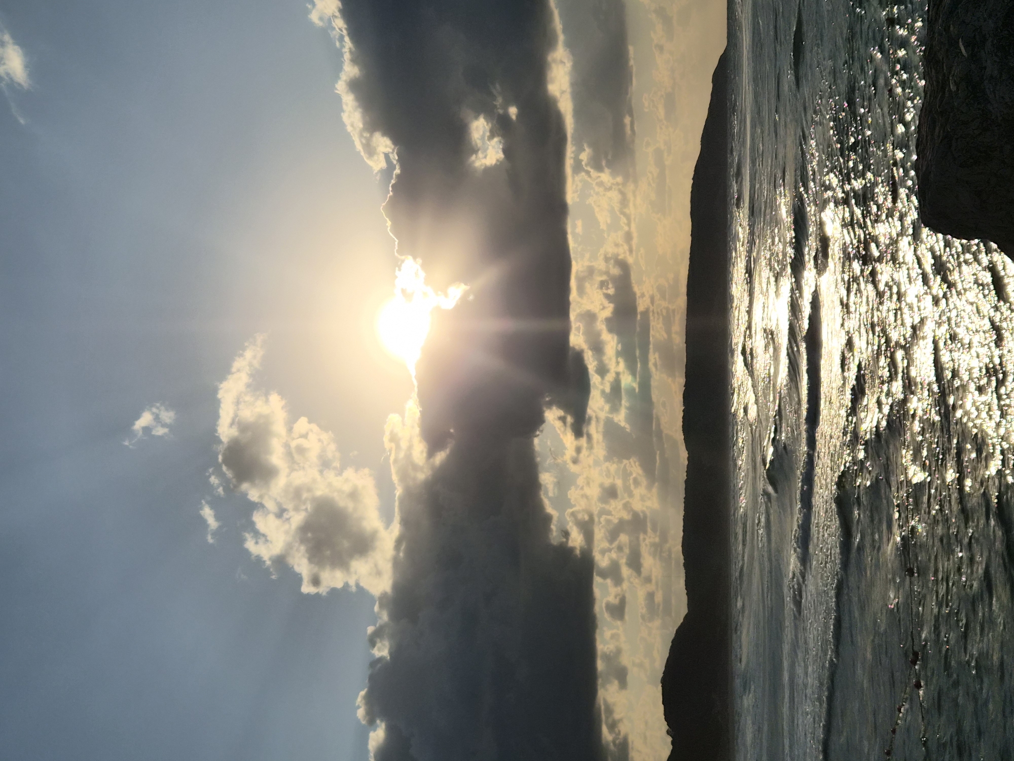The image captures a dramatic sunset over the Black Sea in Şile, Turkey. The sun, partially obscured by dark, dramatic clouds, casts brilliant rays of light across the sky, illuminating the choppy sea below. The water reflects the sunlight in a dazzling display of sparkles and highlights. In the distance, the silhouette of the Şile coast, characterized by rolling hills and a dark, rugged coastline, provides a stark contrast to the luminous sky and sea. The foreground features a dark, rocky outcrop, hinting at the natural beauty of the Şile shore. There are no people or active events depicted in the scene, it is a static, atmospheric landscape.