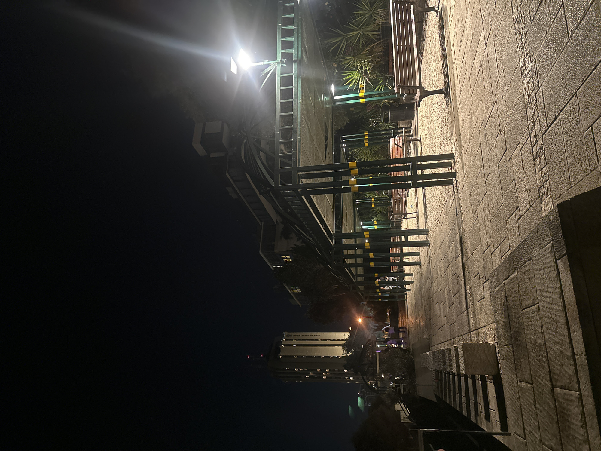 This image captures a nighttime scene on a paved walkway in Haifa, Israel. The walkway is lined with benches and covered seating areas supported by green metal posts with yellow accents. A tall, illuminated building stands in the background, with its windows lit. Several people are visible walking along the pathway. The lighting suggests an urban park or public space. The scene is illuminated by streetlights and the ambient light from the building.