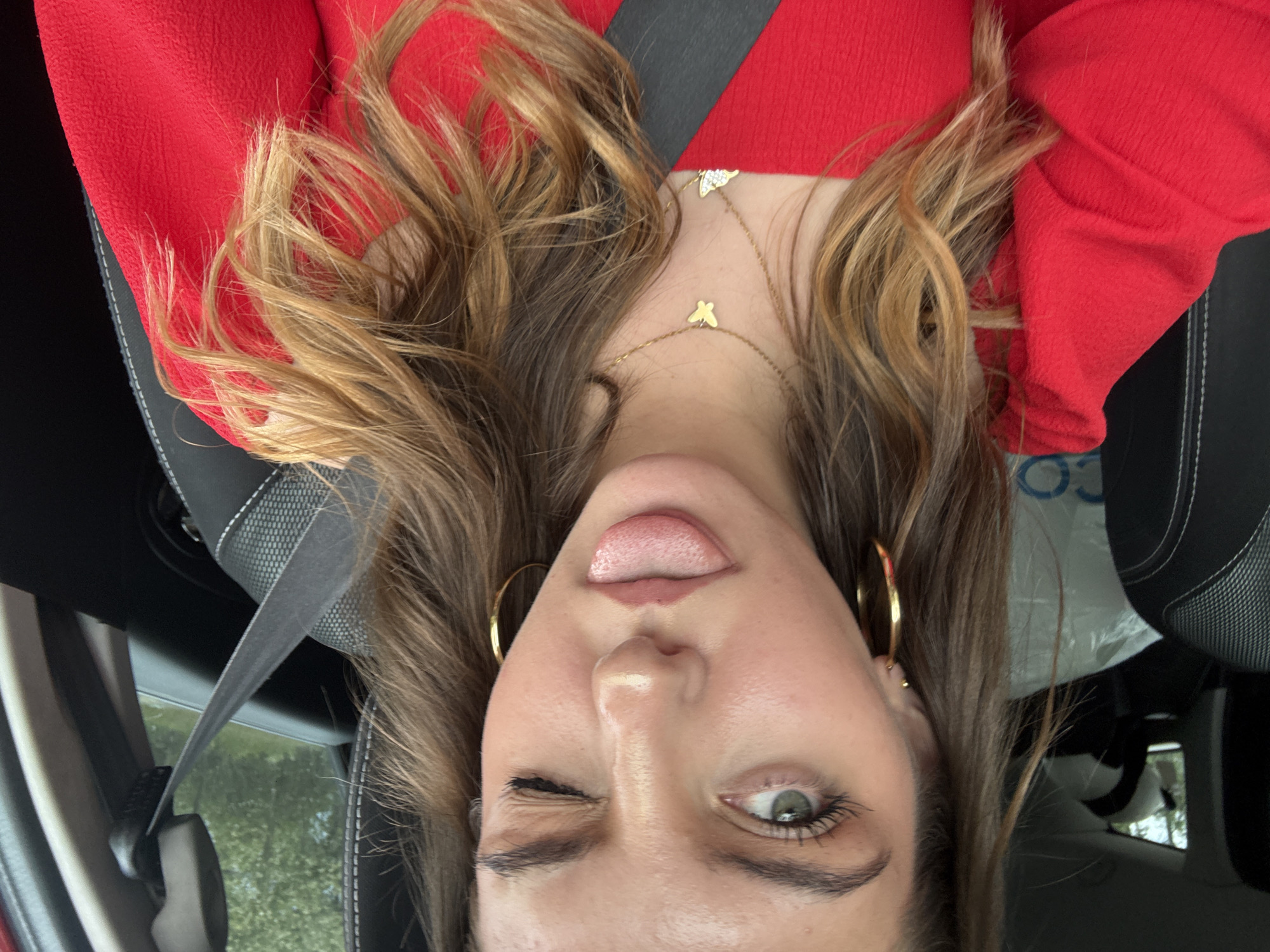 A woman is in the passenger seat of a car, wearing a red shirt and a seatbelt. She has brown hair with blonde highlights, gold hoop earrings, and a gold necklace with multiple pendants. She is sticking her tongue out and winking with her left eye. The location is not evident in the photo, and there is no indication it is in Bliznets, Bulgaria.