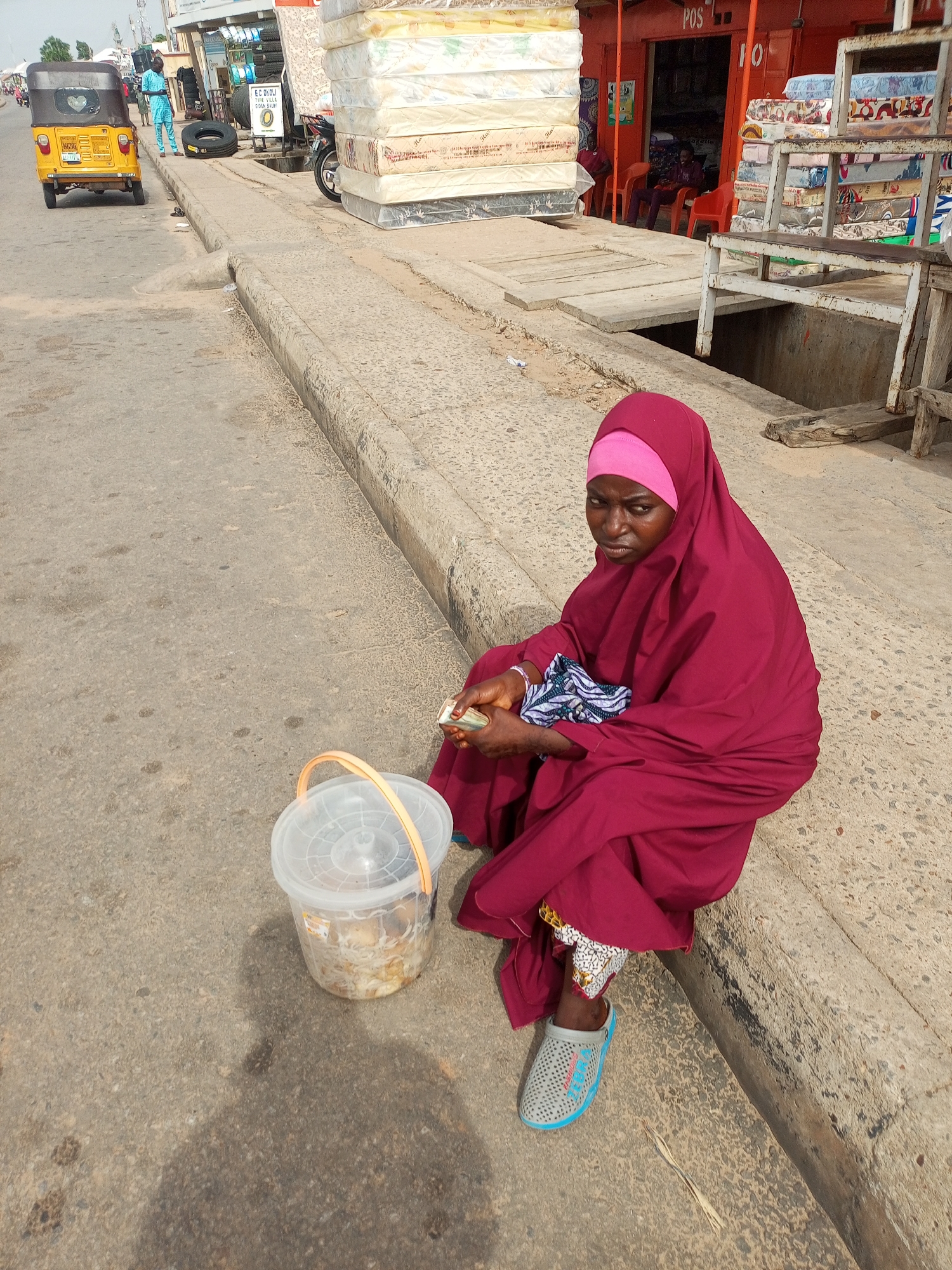 A woman wearing a maroon hijab and dress is sitting on a curb on a paved street. She is holding a small amount of cash in her hands and looking towards the camera. Next to her is a clear plastic bucket with a lid, containing some food items. In the background, there are stacks of mattresses, a yellow tricycle, and buildings. The scene appears to be in an outdoor market or commercial area.