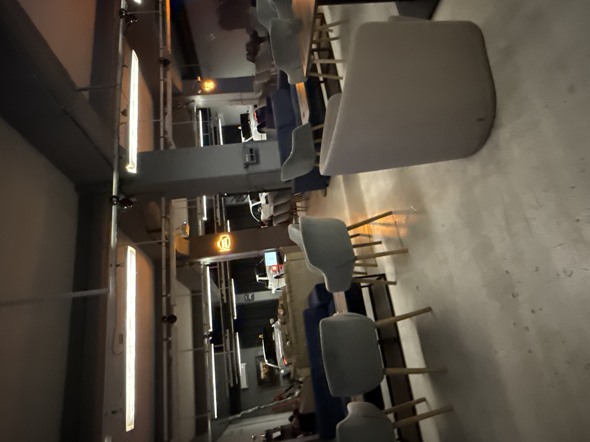 The image depicts an indoor space with several seating areas, suggesting it could be a lounge, cafe, or restaurant. The lighting is dim, with prominent fluorescent tube lights on the ceiling. In the background, white cars are visible, possibly indicating a car showroom, a garage, or a venue that incorporates automotive themes. Some of the cars appear to be part of a display or exhibition. The foreground is dominated by upholstered chairs and sofas in shades of gray and blue, arranged around tables. The overall atmosphere is modern and dimly lit, with a focus on creating a relaxed ambiance. The location of this scene is in the city of Plovdiv, Bulgaria.