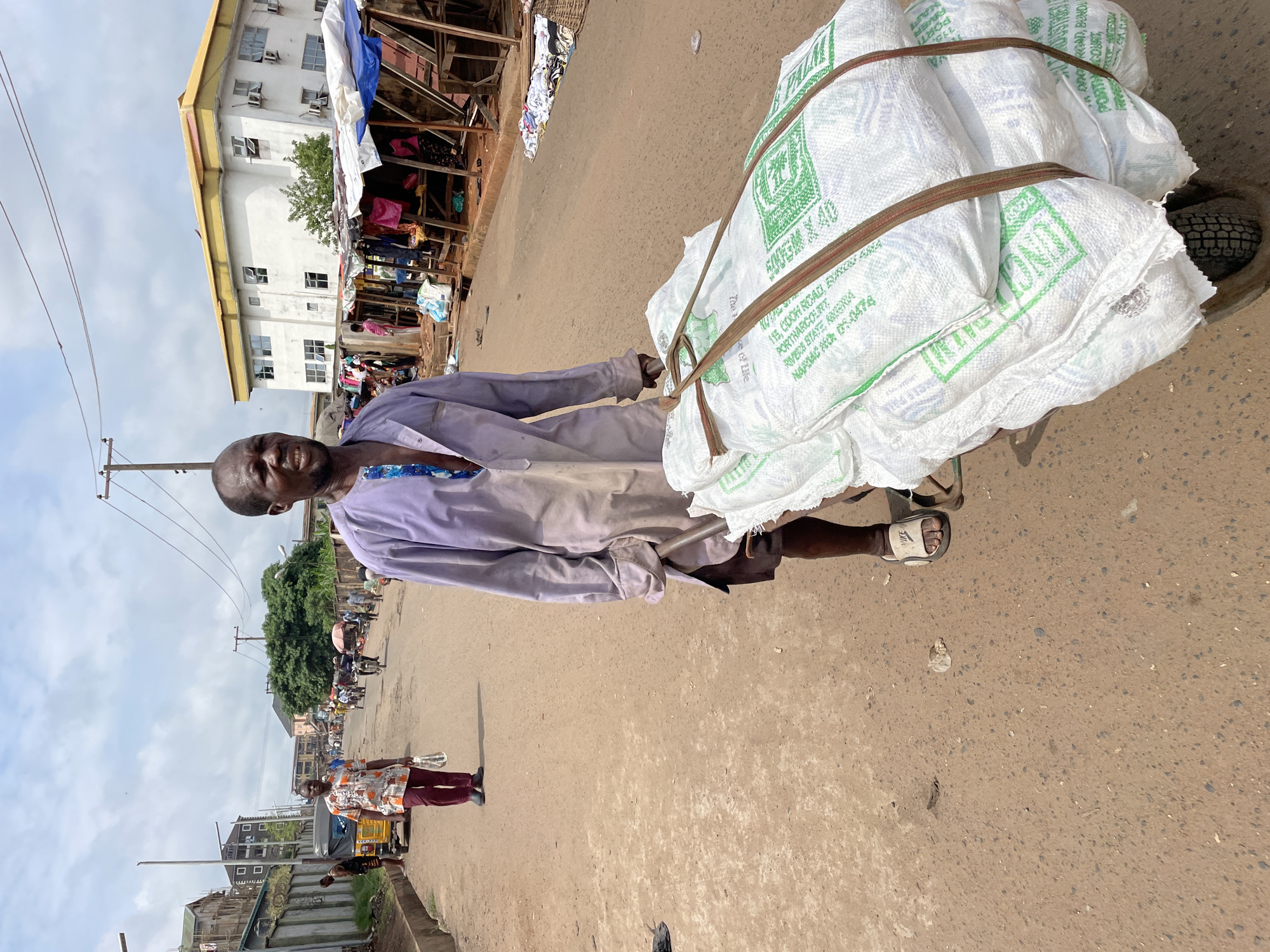 A man pushes a handcart loaded with several large white sacks bound with brown straps. He is wearing a light purple and white gradient shirt and dark shorts, and white slides. The man is walking on a dirt road, with a market on his right. The market consists of wooden stalls with tarpaulin roofs, displaying clothing and other goods. In the background, there are buildings, trees, and a cloudy sky. Another man in an orange and white patterned shirt is walking in the distance on the left side of the road, and a yellow three-wheeled vehicle is parked behind him. There are overhead power lines visible throughout the scene. The sacks on the cart have green text printed on them, including "PORT HARCOURT" and "RIVERS STATE".
