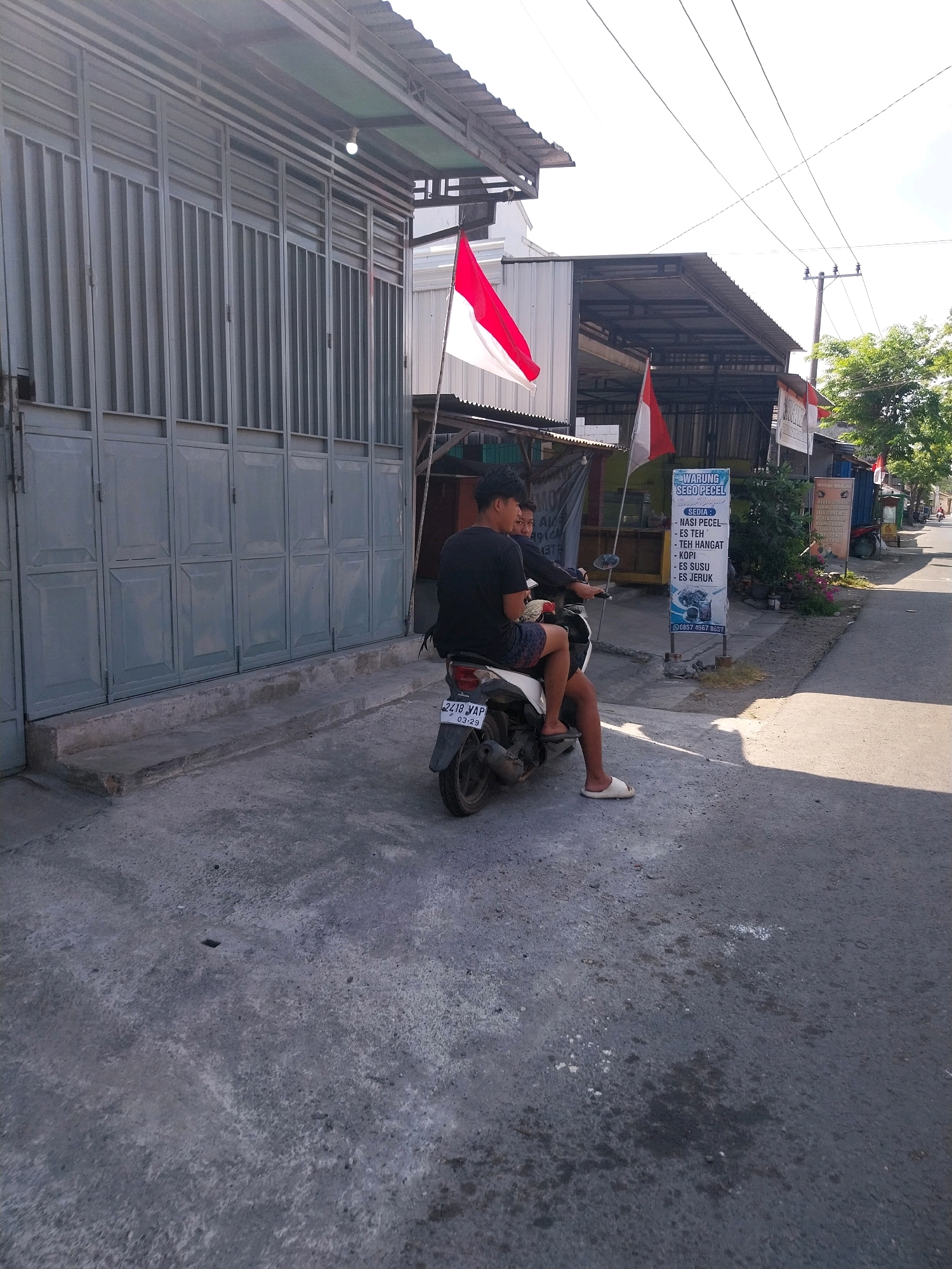 The image shows two young men on a motorbike parked on the side of a street in Campur, Indonesia. The man in front is wearing a black t-shirt and dark shorts, and the man behind him is wearing a black t-shirt and patterned shorts. Both are wearing sandals. The motorbike is a dark color, and its license plate reads "2418 YAP 03-29".
To the left of the motorbike is a building with a gray, corrugated metal facade. To the right of the building, there is a narrow concrete sidewalk, and then the paved street. A sign with Indonesian text, possibly advertising a "WARUNG SEGO PECEL", is visible on a pole by the street. Two Indonesian flags are also displayed on poles near the sign. In the background, there are more buildings, trees, and utility poles, suggesting a semi-urban or rural setting. The sky is bright and appears to be sunny. The overall lighting suggests it is daytime.