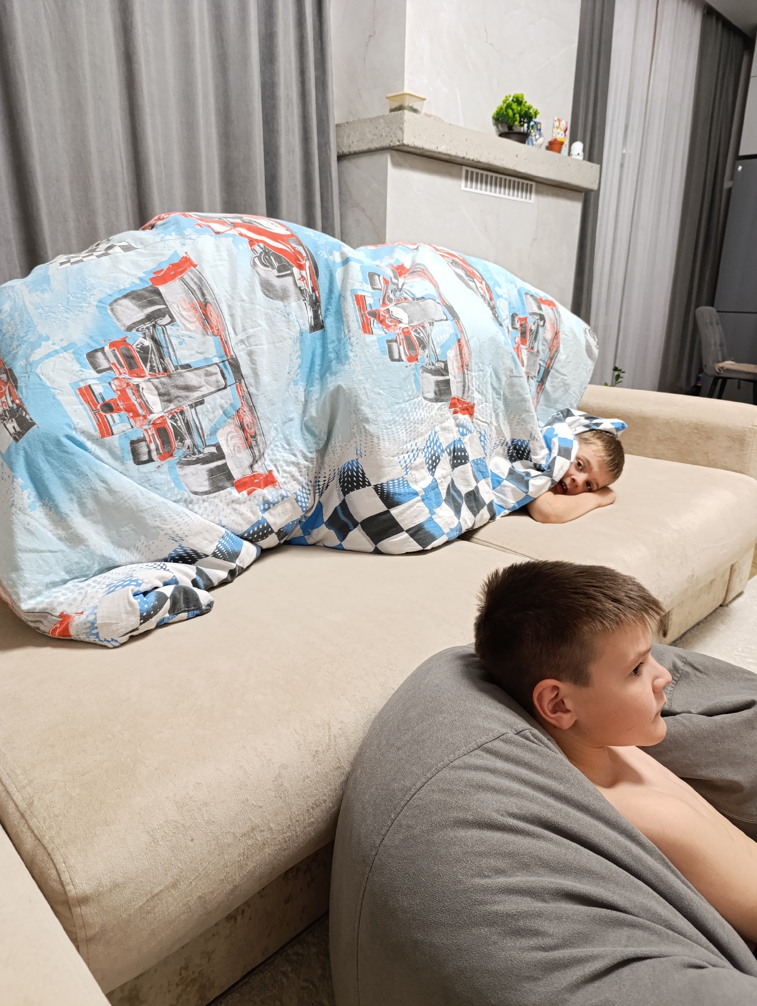 Two young boys are present indoors. One boy is on a beige sofa, partially covered by a blue and white blanket with a race car pattern. He is lying down, with his head near the edge of the sofa. The second boy is seated in a gray beanbag chair in the foreground. He is shirtless and looking off to the side.
In the background, there is a decorative mantelpiece with small objects on top. Light gray curtains are visible near a wall. The setting appears to be a home interior. The city of Chaikovsky, Russia, is not visually confirmed in the image.
