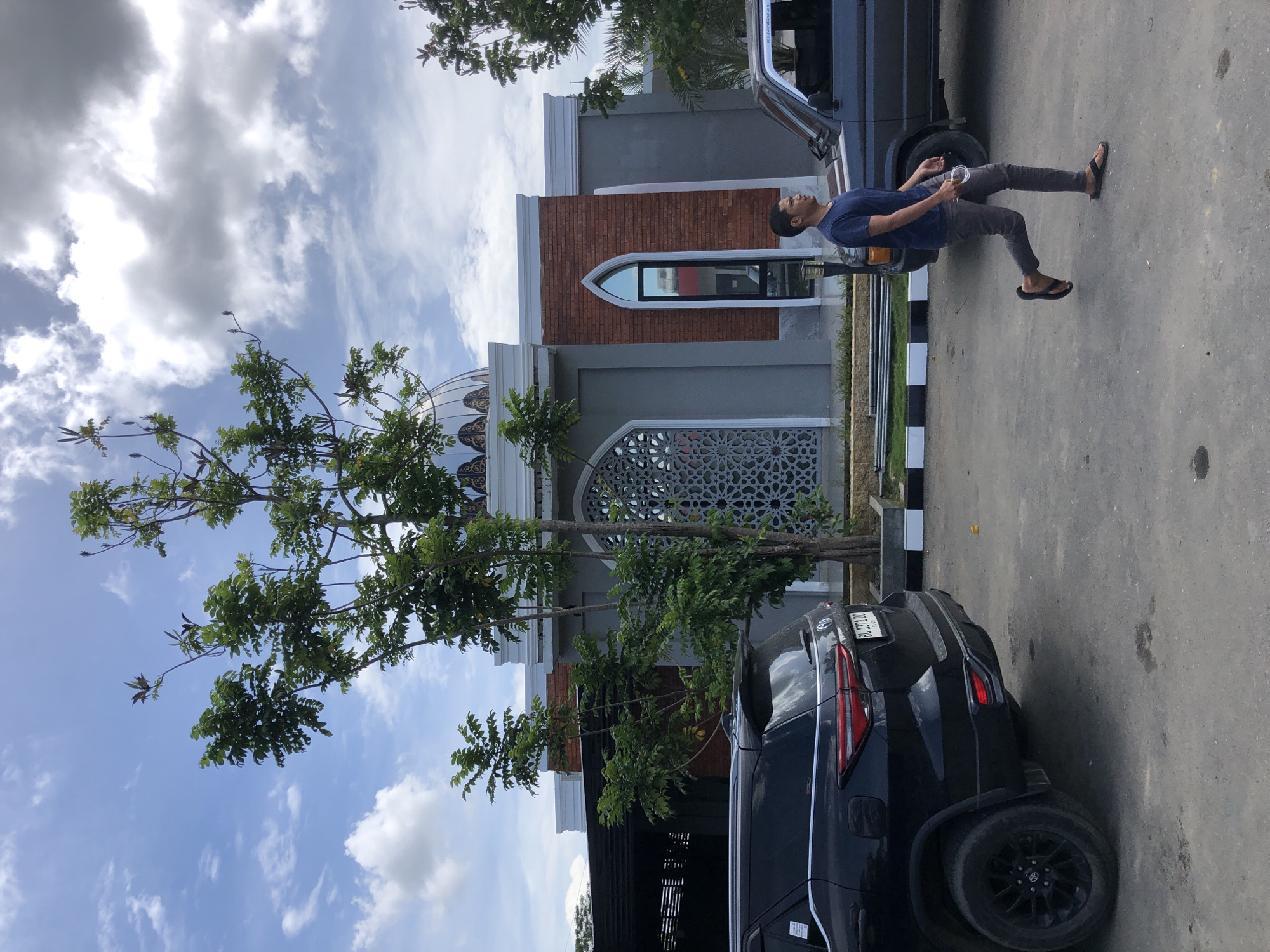 The image displays an outdoor daytime scene in Lhoksukon, Indonesia, featuring one male individual, two vehicles, and a building under a partly cloudy sky.
A light gray building with white trim, possibly a mosque due to its dome structure, occupies the background. The dome is light-colored with dark, intricate patterns. The building's facade includes a red brick section with an arched window on the right and a large arched opening with an ornate geometric screen on the left.
In the mid-ground, a male subject with short dark hair, wearing a dark blue t-shirt, dark gray pants, and black flip-flops, is walking from right to left. He holds a white object, possibly a phone with a lanyard or cable, in his left hand.
In the left foreground, the rear of a black Toyota Fortuner SUV is visible, displaying the license plate "BL 1371 DO". Its rear wheels are black multi-spoke rims. In the right mid-ground, partially obscured by the walking man, is the front passenger side of an older model, dark gray SUV, featuring an orange indicator light on its bumper.
The ground is a worn, light gray asphalt or concrete surface. A low curb with alternating black and white stripes separates the paved area from a landscaped strip adjacent to the building. Several trees with green foliage are present, with one prominent tree positioned centrally in front of the building. The sky overhead is bright blue with scattered white and gray clouds.