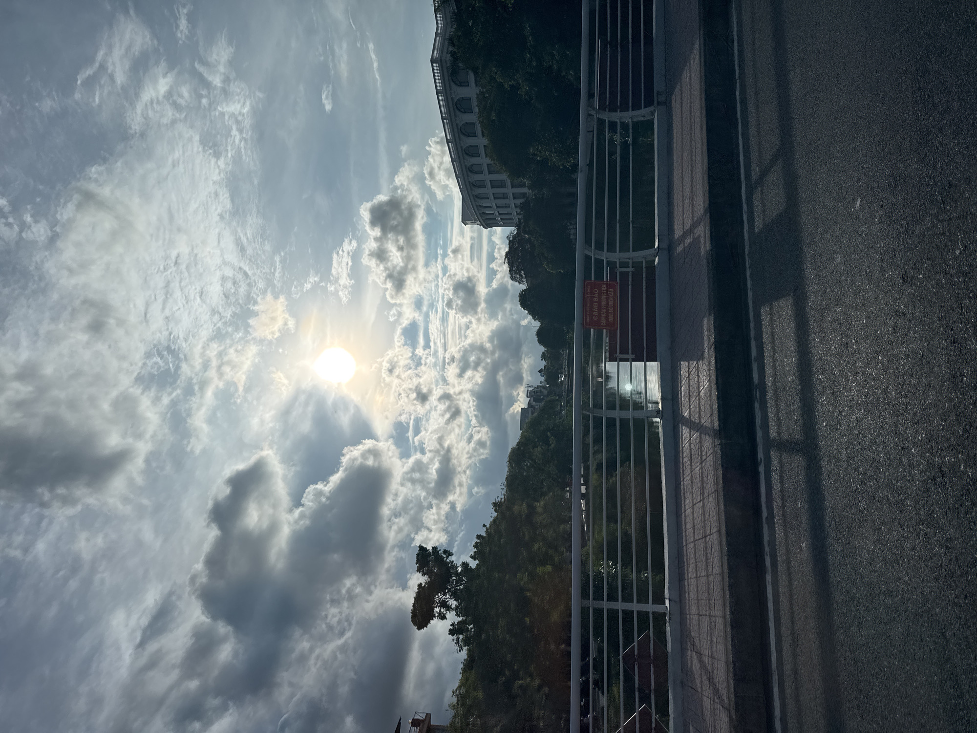 The image depicts a bright, overcast day in Thuận Hóa Ward, Vietnam, viewed from a road that appears to be elevated, possibly a bridge or overpass. The sun is visible through the clouds, casting a bright glow. Below the road is a body of water, with lush green trees lining its banks. On the far side of the water, a large, multi-story building with arched windows is visible, surrounded by more trees. A white metal railing runs along the edge of the road, with a red sign attached to it. The road itself is paved, with shadows from the railing cast upon it. The overall impression is one of a serene, daytime scene in an urban environment, with a blend of natural and architectural elements.