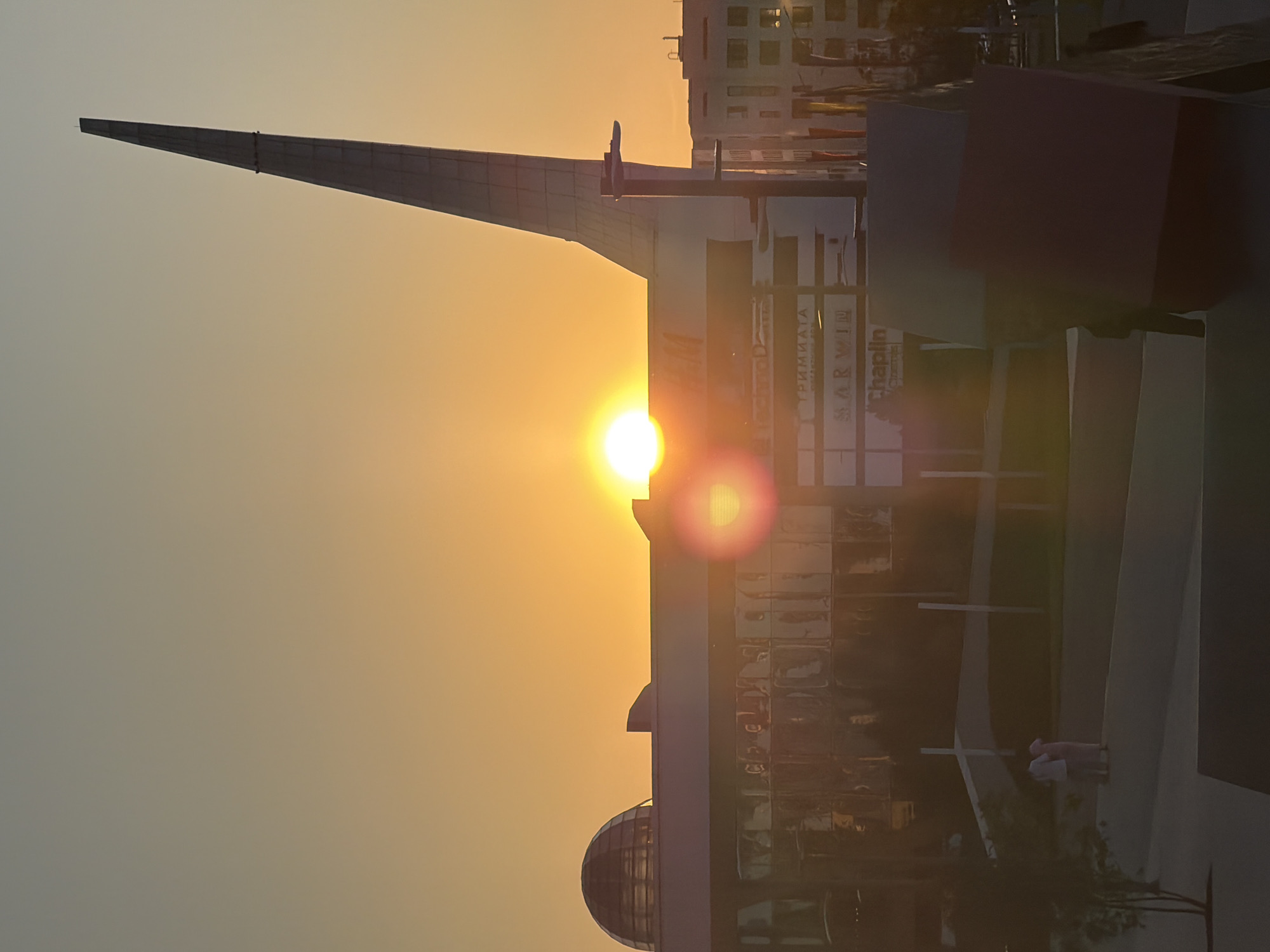 This image captures a sunset over a modern urban landscape in Astana, Kazakhstan. The sun, a bright orb, is positioned near the horizon, casting a warm orange glow across the sky and reflecting off the glass facade of a large building.
Dominating the skyline is a tall, slender, pointed structure, possibly a monument or part of a skyscraper. Adjacent to it, a building with a domed roof is visible. The main building features large glass panels, creating a mirrored effect that reflects the surrounding environment and the sunset. Signs are visible on the building, including text that appears to be in Cyrillic script. One sign reads "TRIMMIATA," another "MARWIN," and below that, "Chaplin Cinema."
In the foreground, a few individuals are present, appearing to walk or stand in a paved area. The scene suggests an outdoor public space, possibly a plaza or walkway in front of the buildings. The overall impression is one of contemporary architecture set against the dramatic backdrop of a sunset.