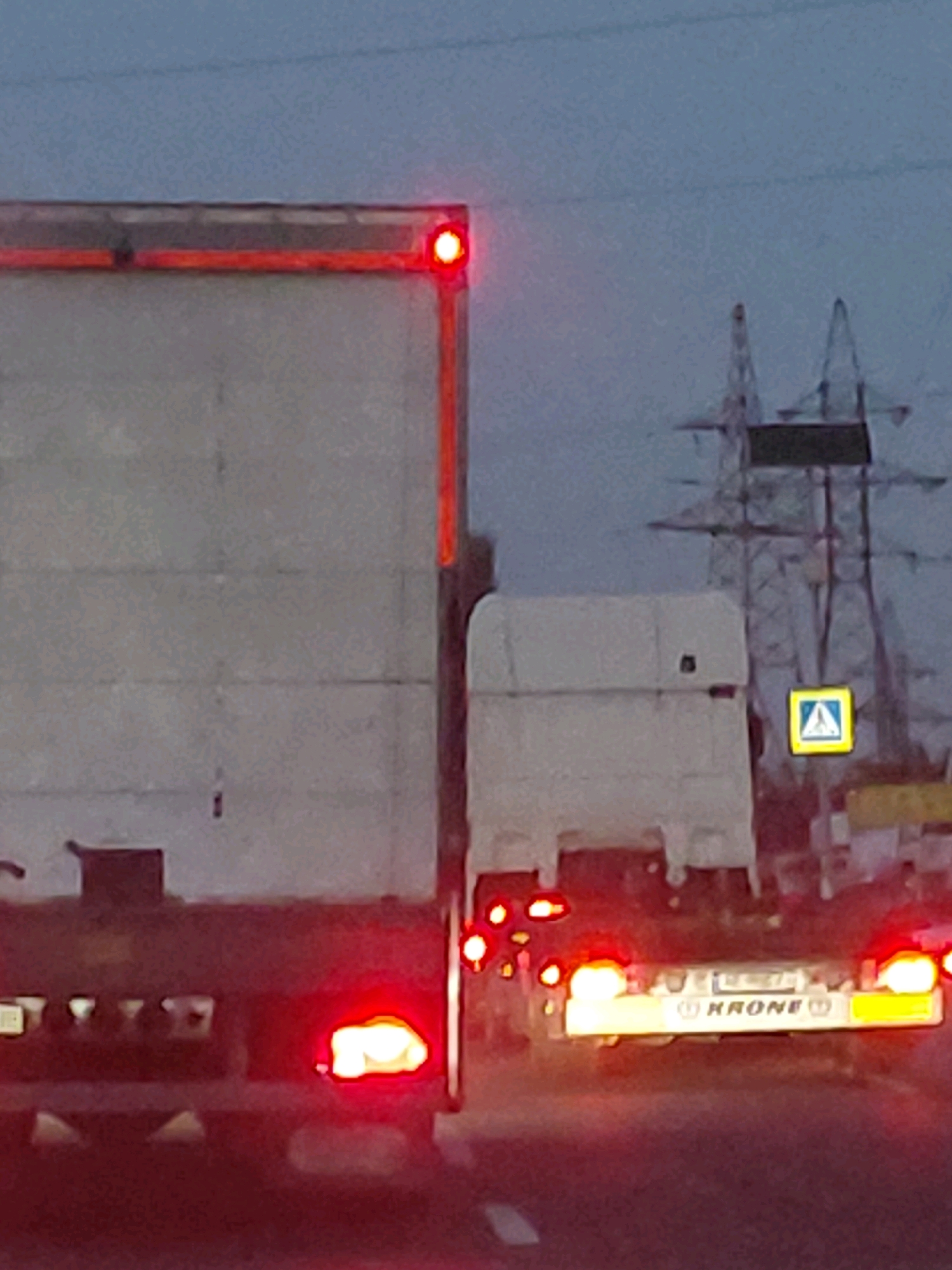 The image captures a close-up, ground-level, eye-level, horizontal shot of a traffic scene at dusk. The primary subjects are the rear ends of two large trucks. The truck on the left is larger and partially obscures the truck on the right. Both trucks have their brake lights illuminated, casting a red glow. The truck on the right displays a white license plate with some illegible text and a prominent yellow and white "KRONE" logo. A pedestrian crossing sign, a yellow square with a blue border and a white symbol of a person walking, is visible to the right of the trucks. In the background, large power lines and pylons dominate the skyline against a dusky sky. The overall impression is one of traffic congestion or a pause in movement during twilight.