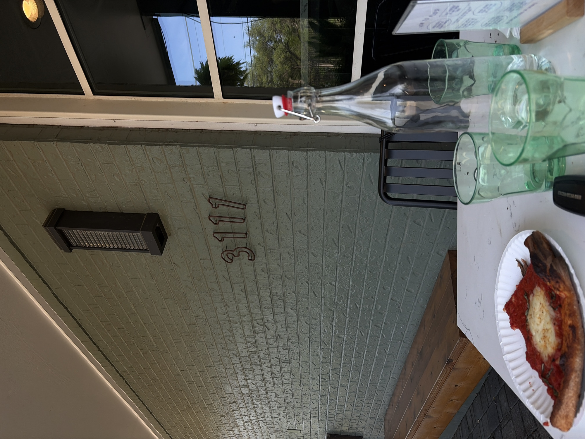 Here is a description of the image:
The image shows the facade of a building with the address "3117". There is a window above the address and a light fixture to the left of the numbers. To the right, there is a black metal chair and a table. On the table, there is a glass bottle, several green glasses, and a plate with a slice of pizza on it. The building has a brick facade and a light-colored roof. There is a wooden beam visible as well.
Based on the address visible, the location is likely in Austin, Texas, United States of America.