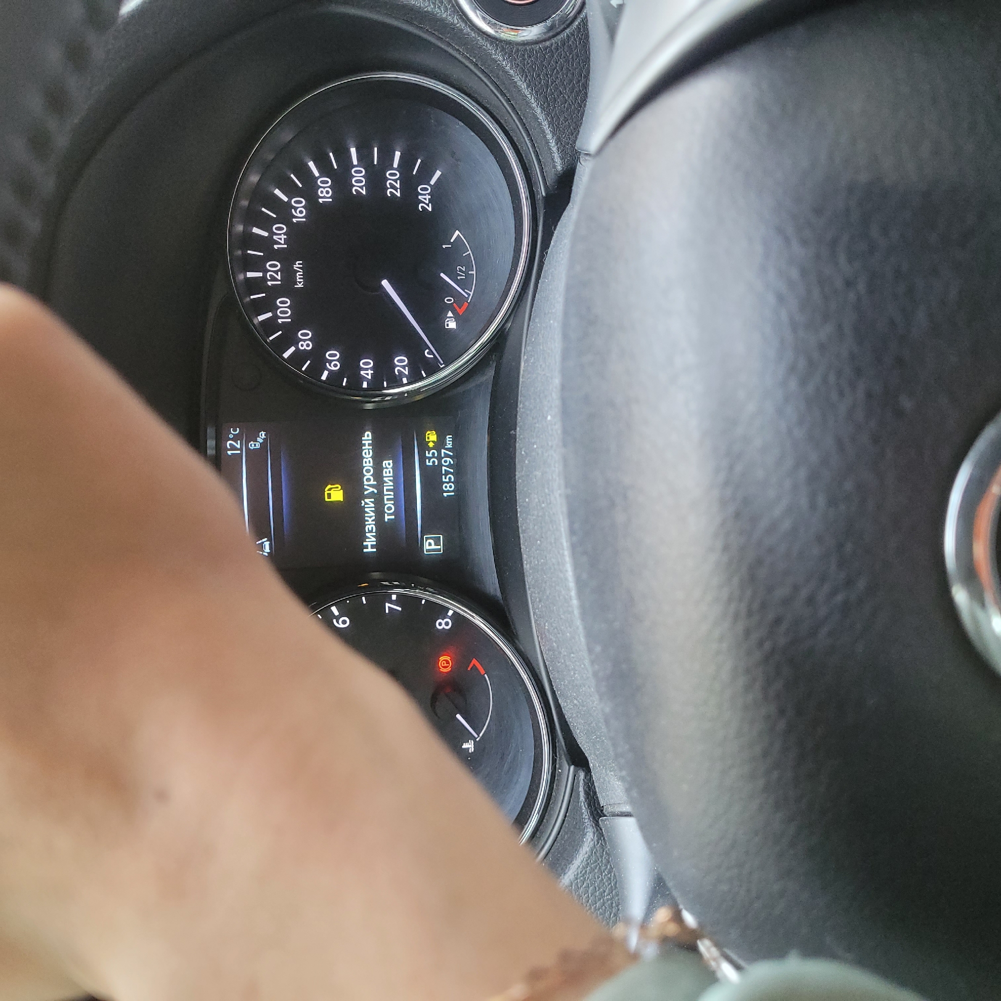 The image shows a close-up view of a car's dashboard, with a hand partially obscuring the upper portion of the frame. The dashboard displays a speedometer, a fuel gauge, and a temperature reading. The speedometer shows a range from 0 to 240 km/h, with the needle indicating a speed below 40 km/h. The fuel gauge is on empty, and a yellow fuel can icon is illuminated, accompanied by Russian text that reads "Низкий уровень топлива" (Low fuel level). The temperature gauge shows 12°C. There is also a "P" symbol illuminated, indicating the car is in park. The odometer reads 185797 km. The overall environment appears to be inside a vehicle, likely in motion or recently stopped. No specific city or location details are discernible from the image.