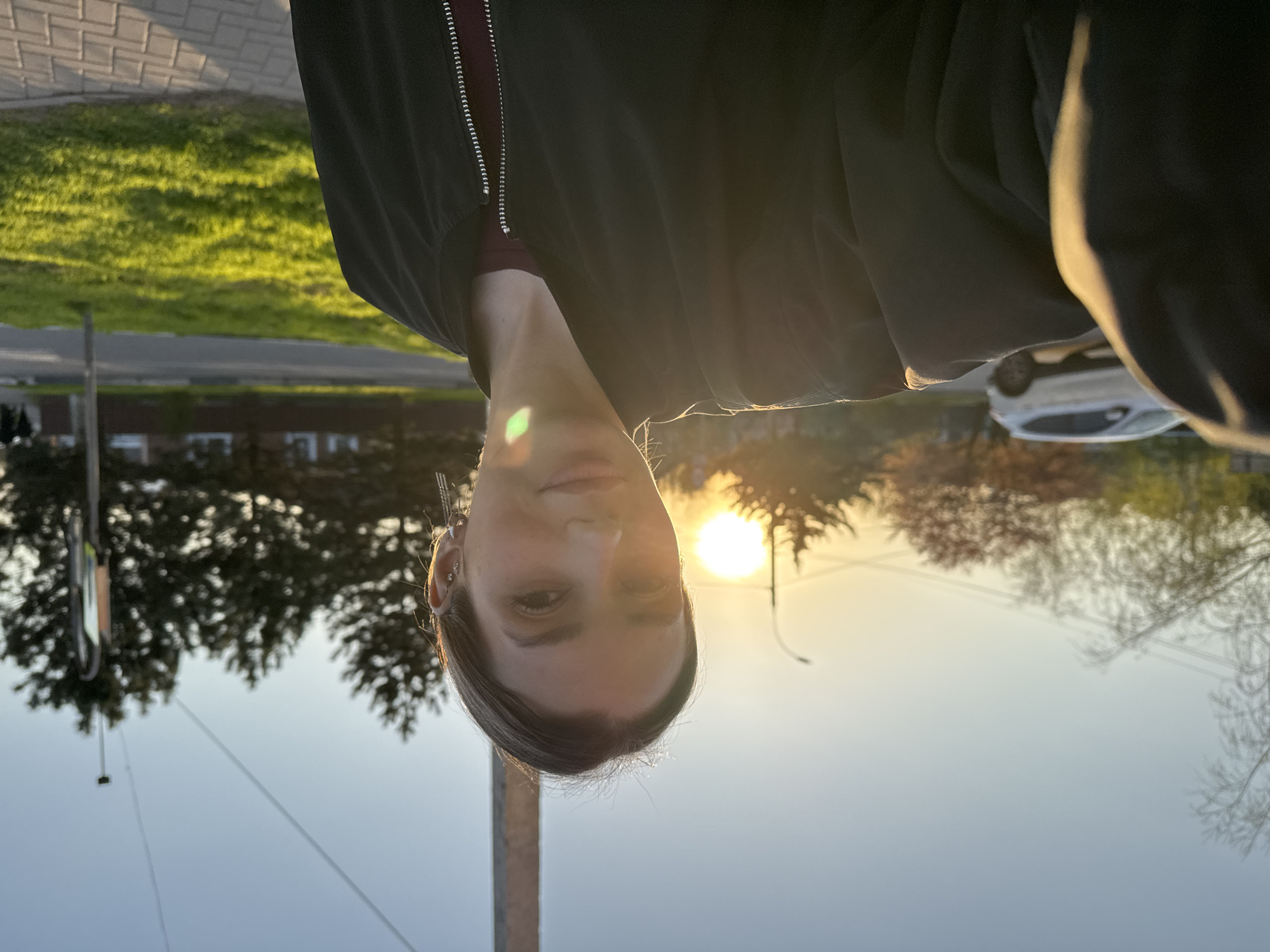 A person is facing the camera with a dark jacket and a red undershirt, and the sun is behind the person. The location appears to be in a city with trees and buildings in the background. There is a car and a light pole visible in the background. The photograph is oriented upside down.